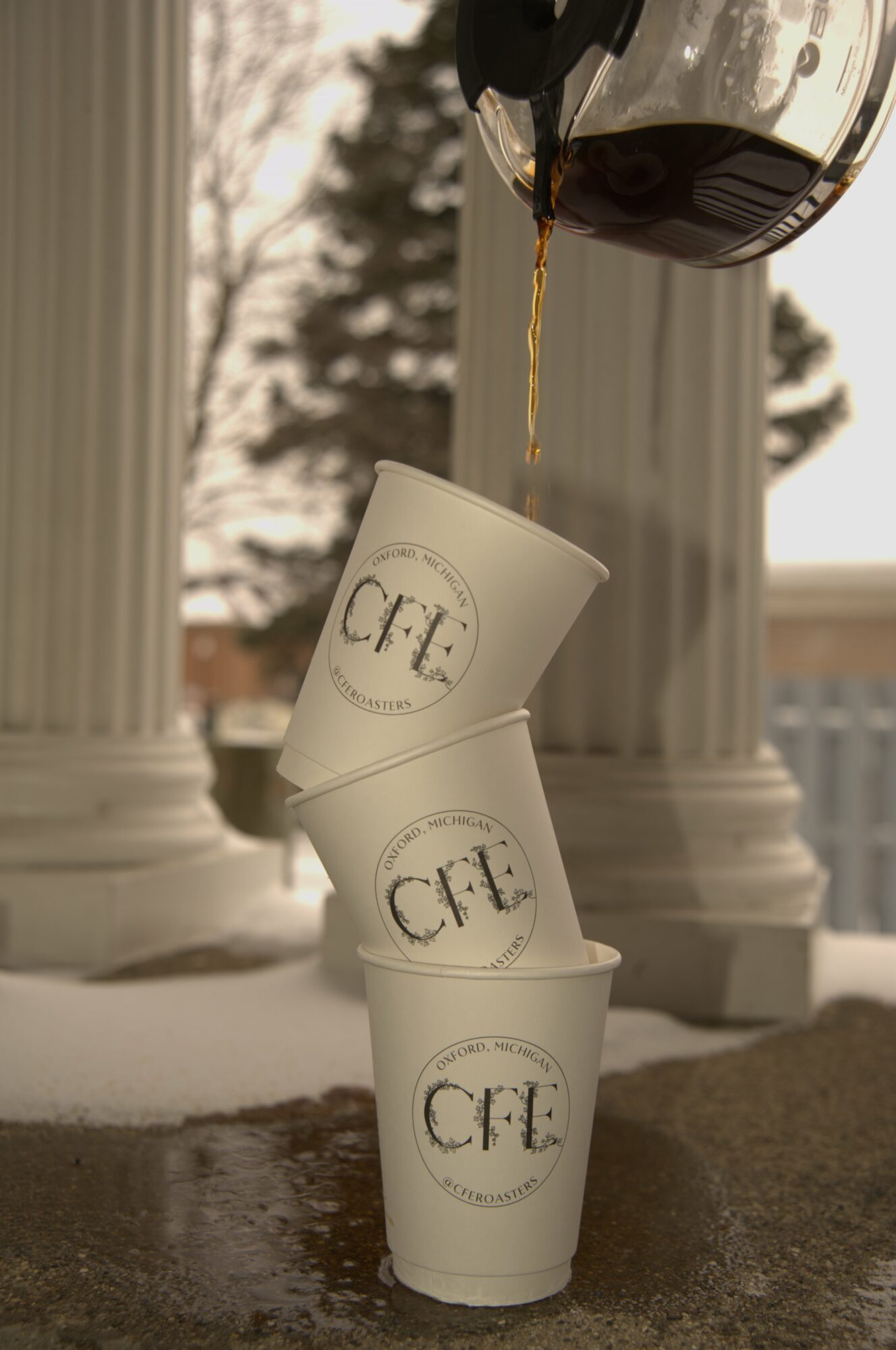 Coffee cups stacked with coffee being poured from a pot into the top cup, outdoors with columns and trees in background.