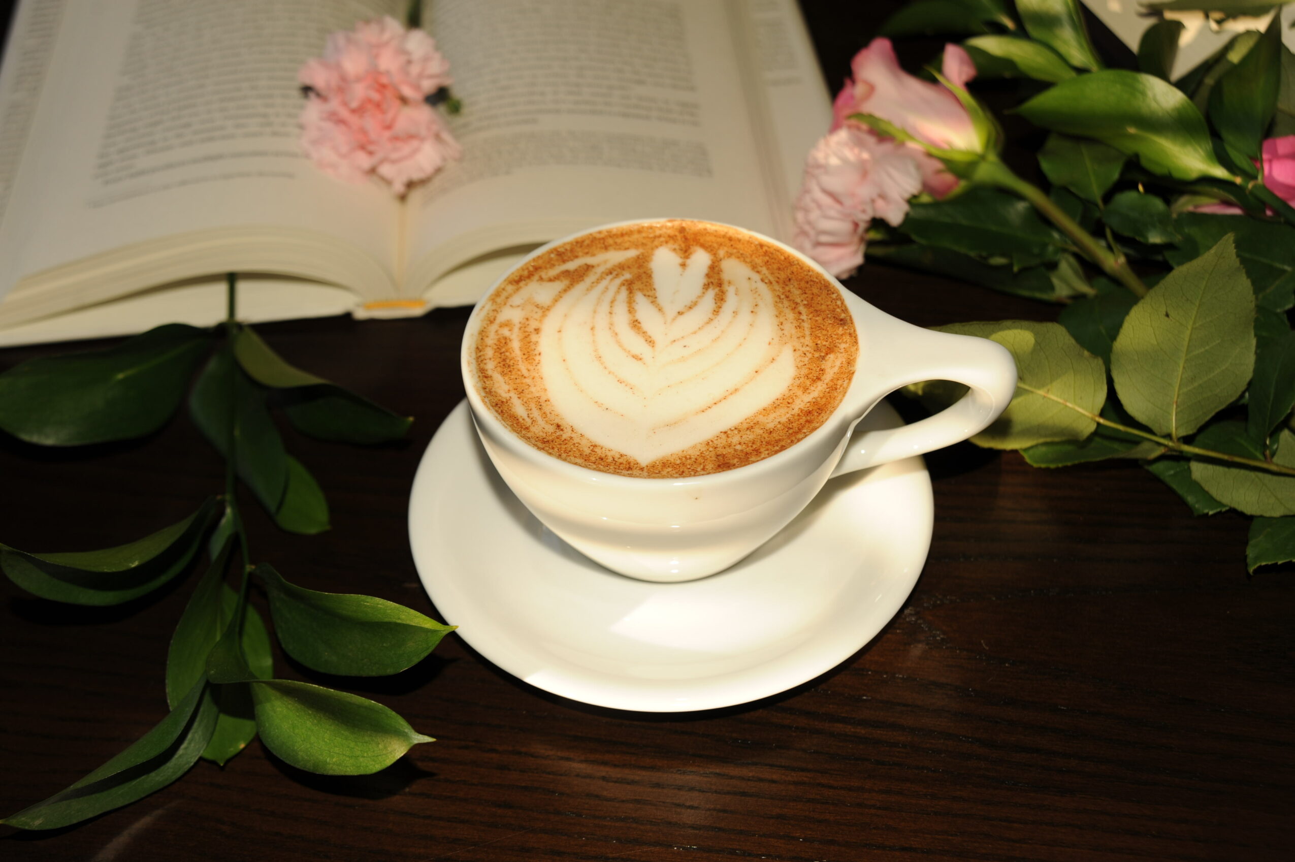 Cup of coffee with latte art on a saucer, surrounded by pink flowers and green leaves, with an open book in background.