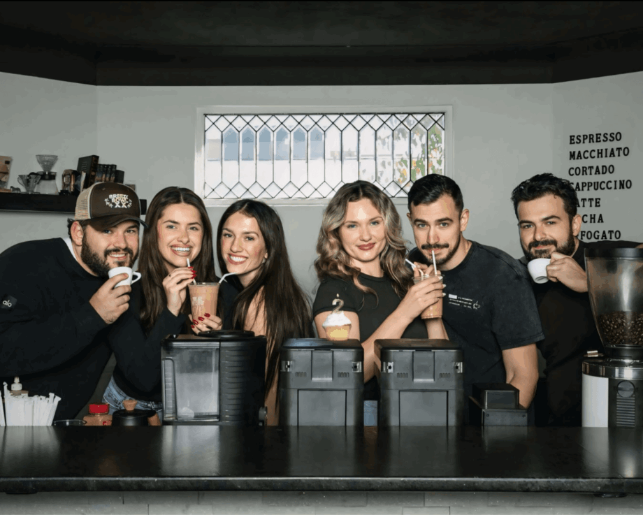 Group of six people behind a coffee bar, smiling and posing for the photo.