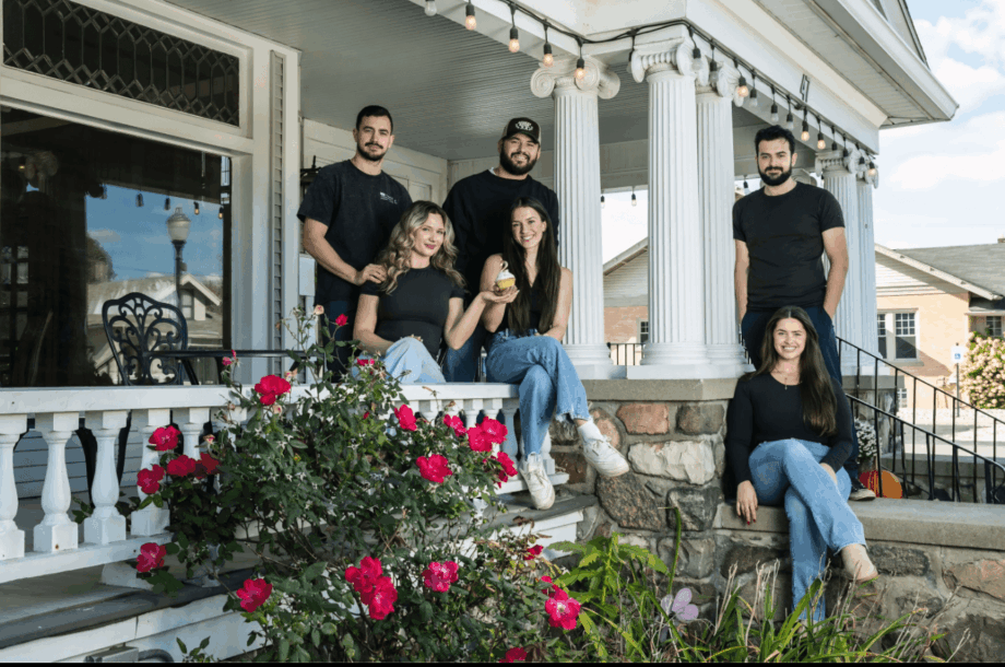 Group of six people sitting and standing on porch with flowers and house in background.