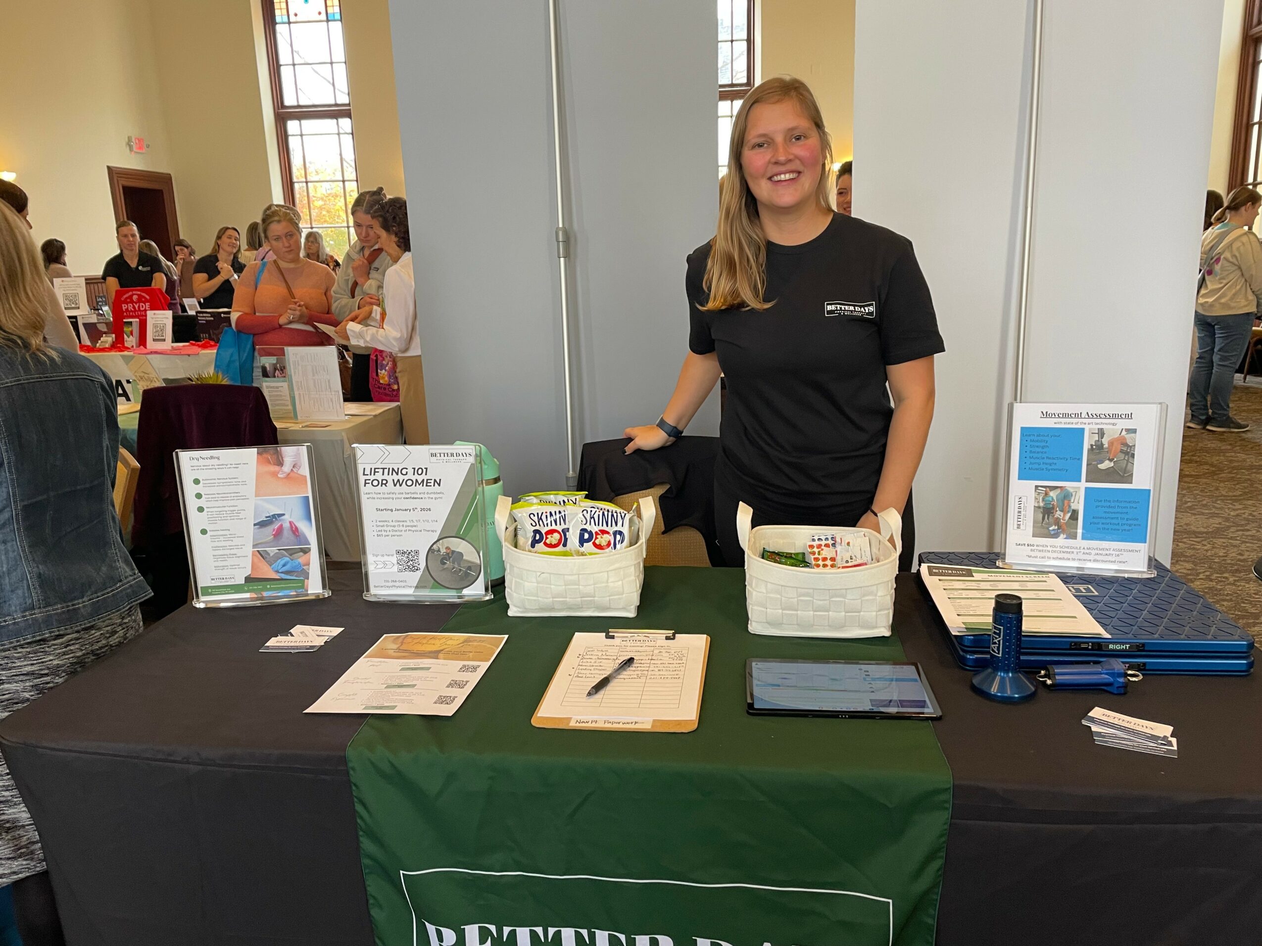 Smiling woman standing behind a table with informational materials at an indoor event, with other people in the background.