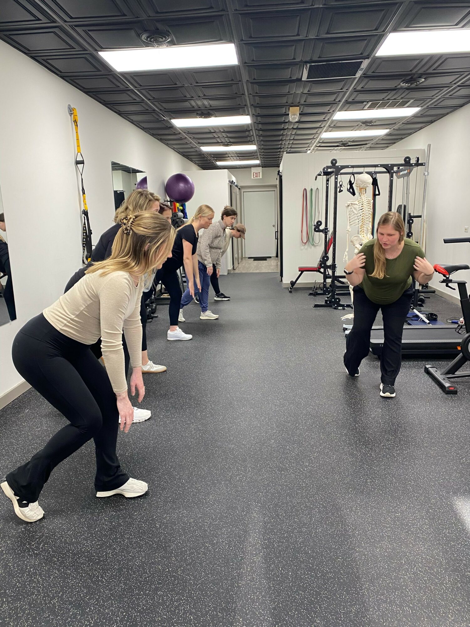 Four women in a gym, one stretching on the floor, others standing and talking, gym equipment in background.