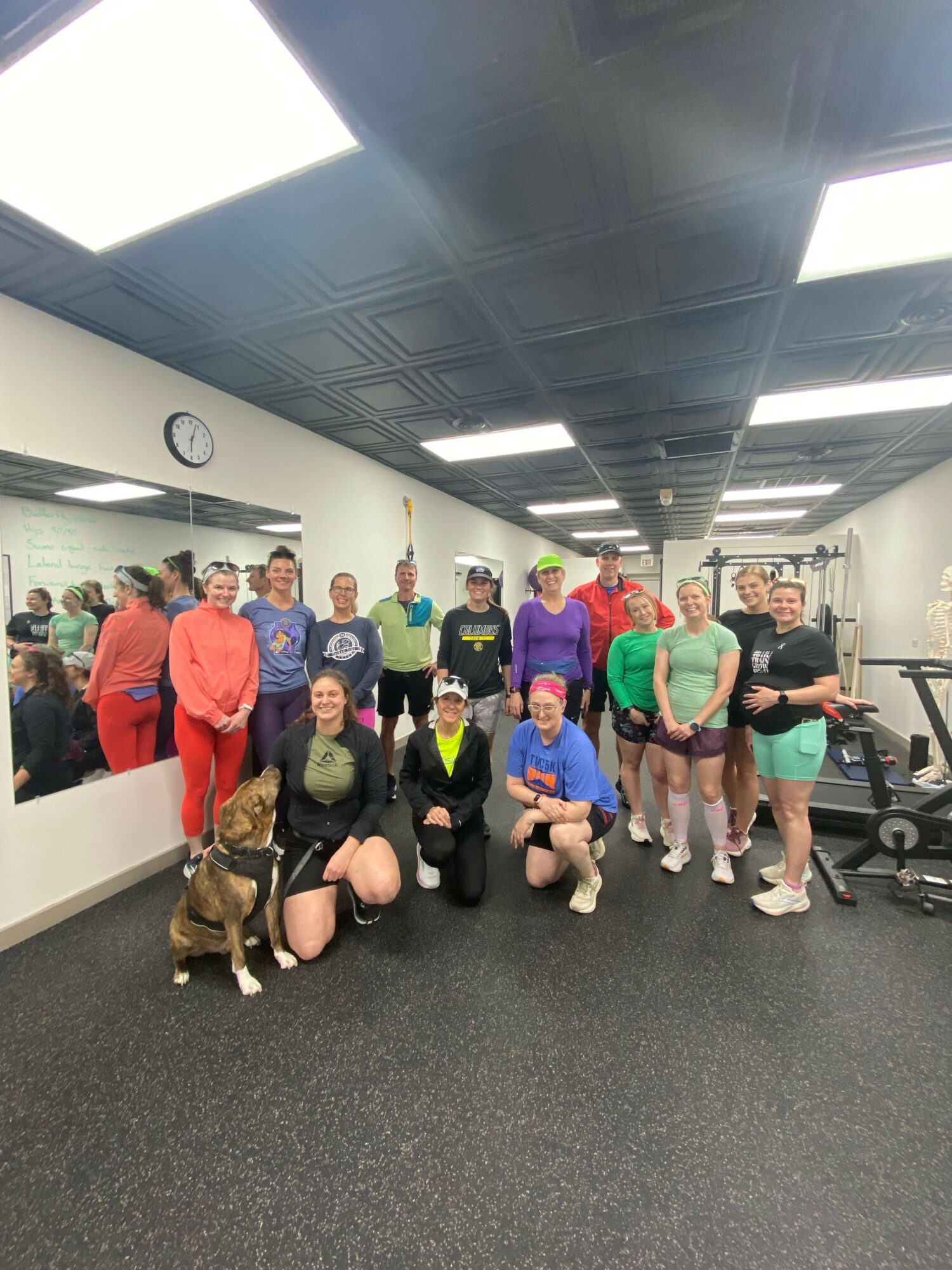 Group of people in a gym, some sitting and some standing, with gym equipment and a clock on the wall.