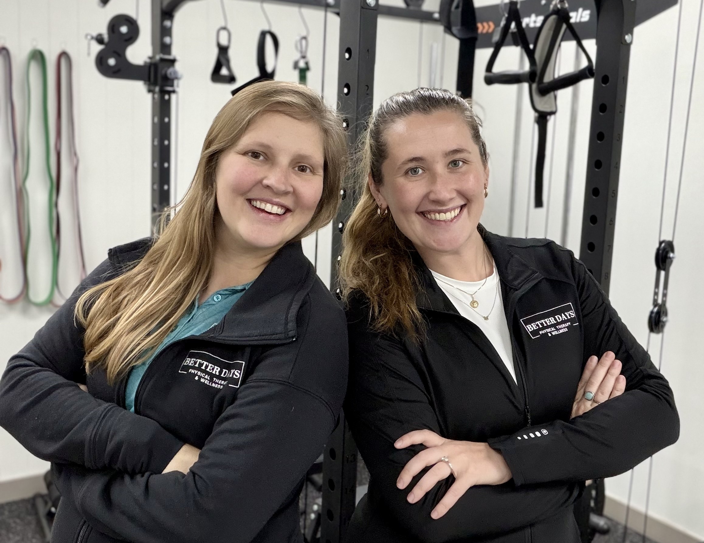 Two women smiling with arms crossed, standing in a gym with workout equipment behind them.