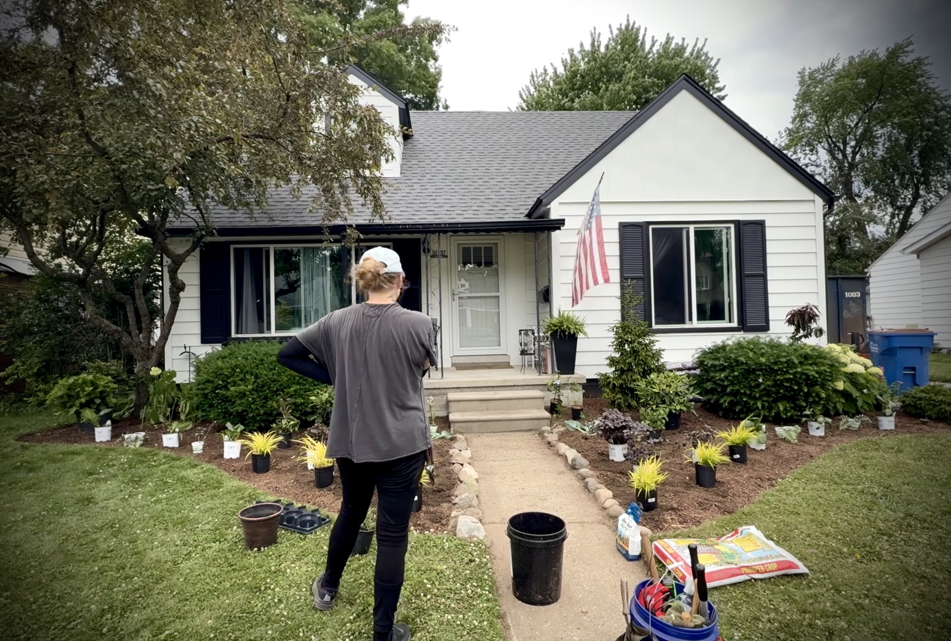 Person standing in front of a white house with a garden, American flag, and gardening supplies nearby.
