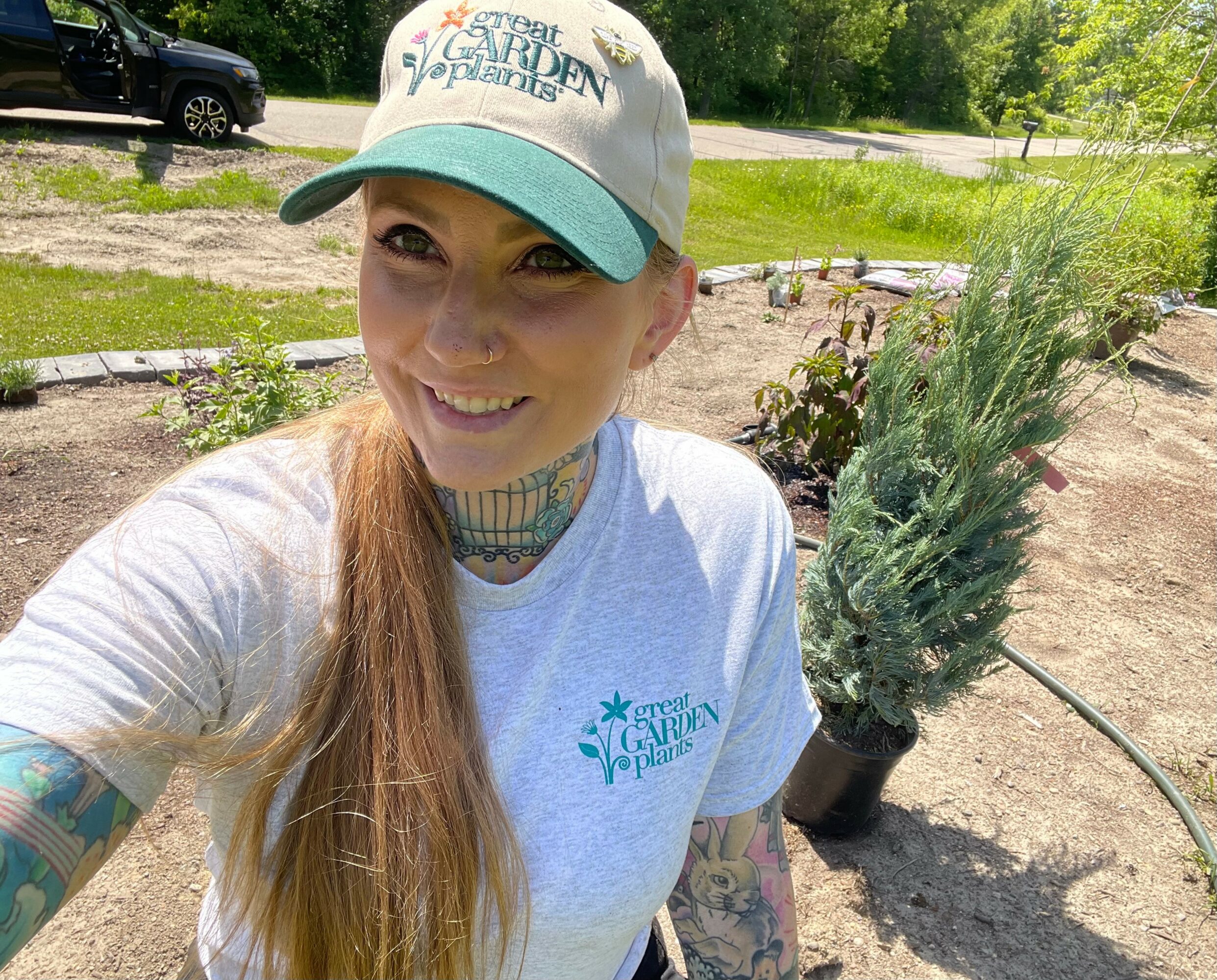 Woman with long hair, wearing a cap and white shirt, smiling outdoors near plants and garden bed.