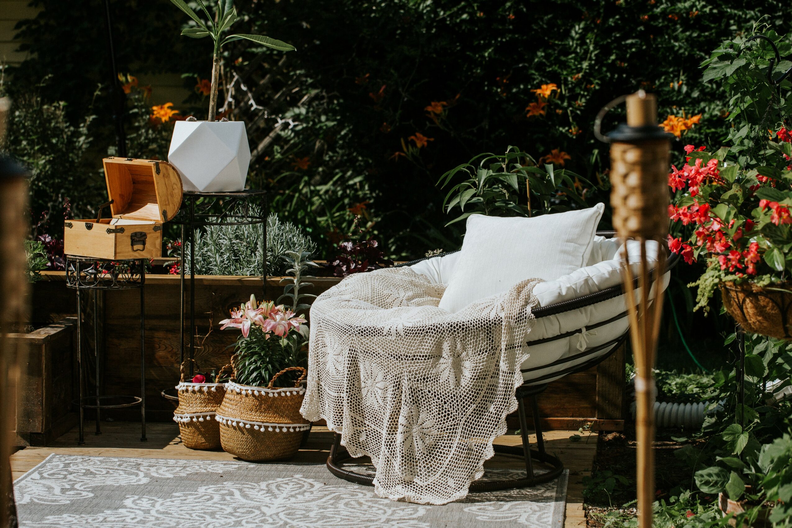 Outdoor scene with a lounge chair, white pillow, lace blanket, potted plants, and flowers, surrounded by greenery.
