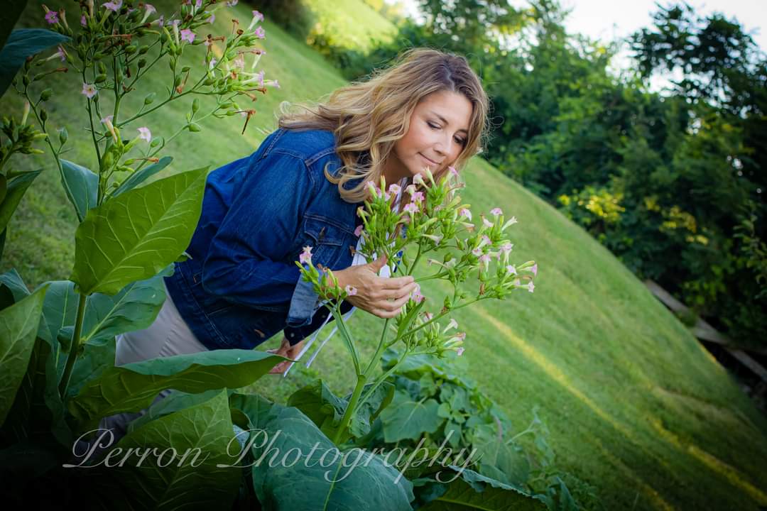 Woman with curly hair in a blue jacket smelling pink flowers in a garden, green grass and trees in background.