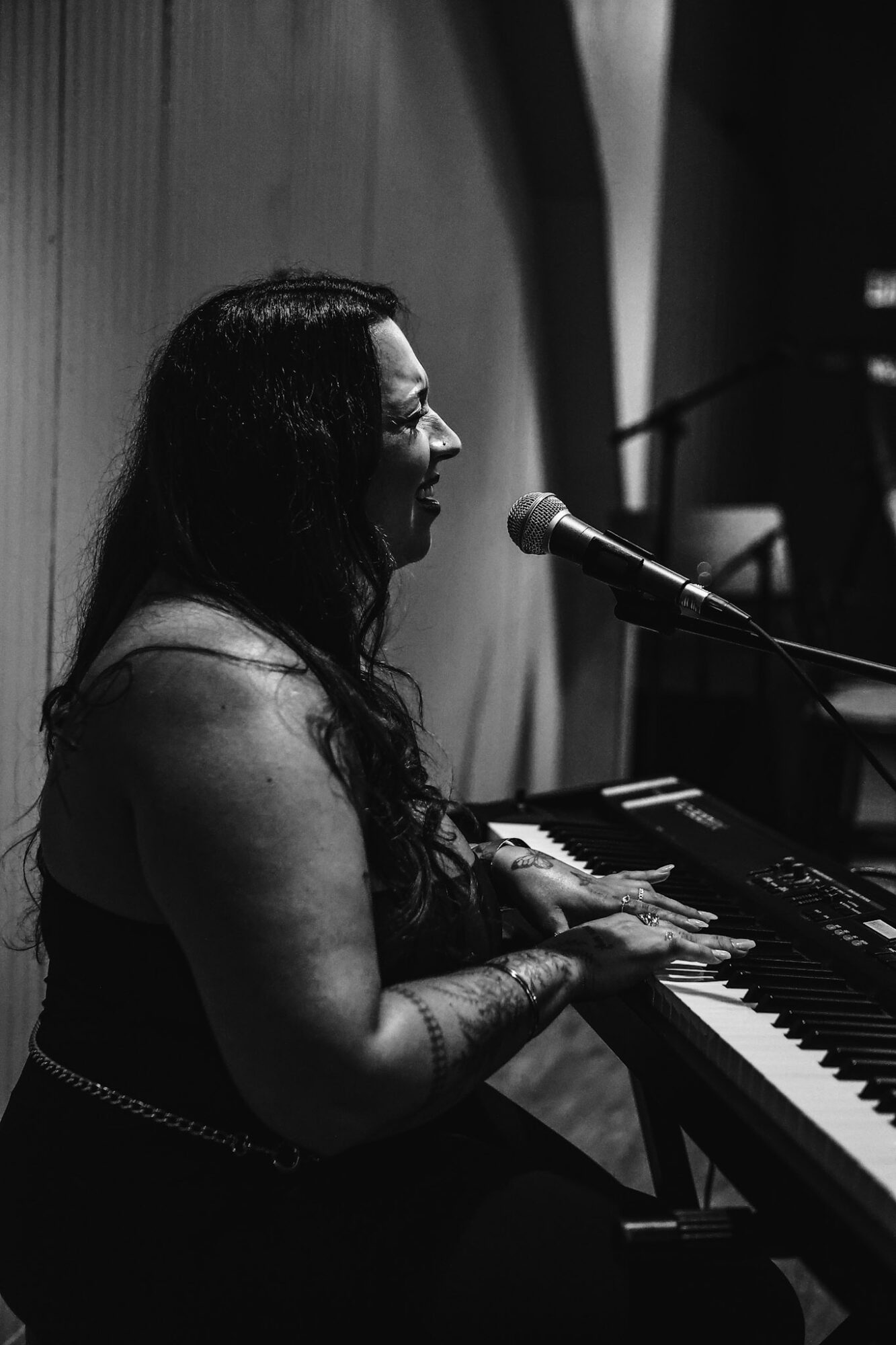Woman with long dark hair playing keyboard and singing into a microphone in a dimly lit room.