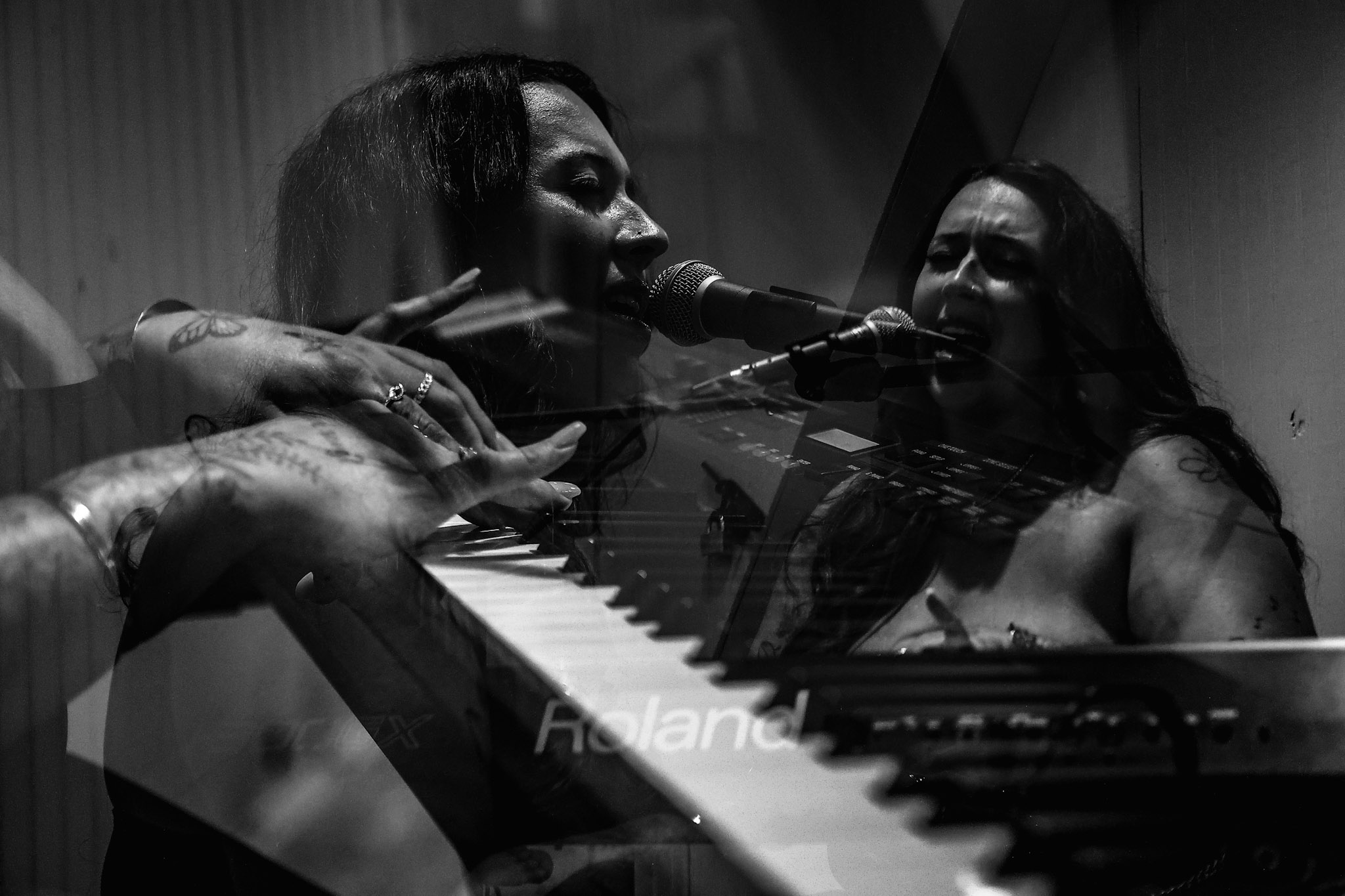 Two women singing into microphones, one playing piano, in a black and white setting.