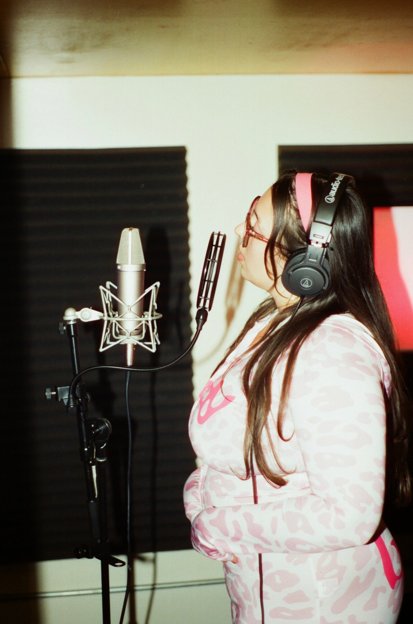 Woman with long hair wearing headphones and glasses, singing into a microphone in a recording studio.