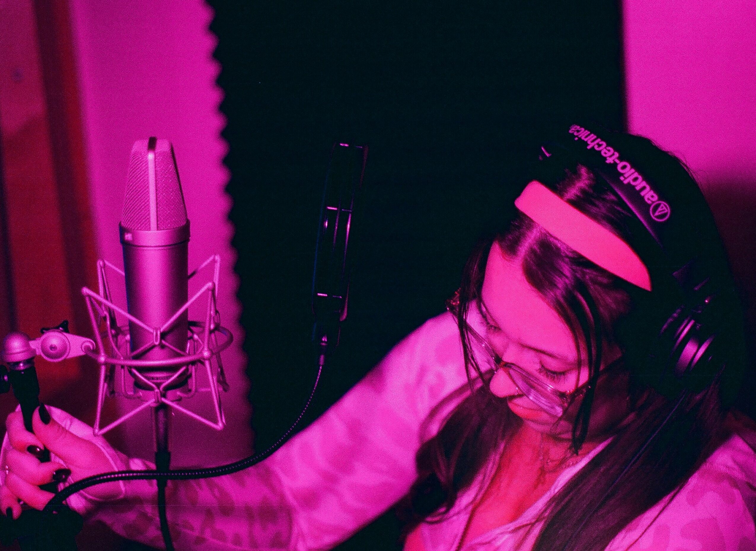 Person wearing headphones and glasses, speaking into a microphone in a recording studio, illuminated by pink lighting.