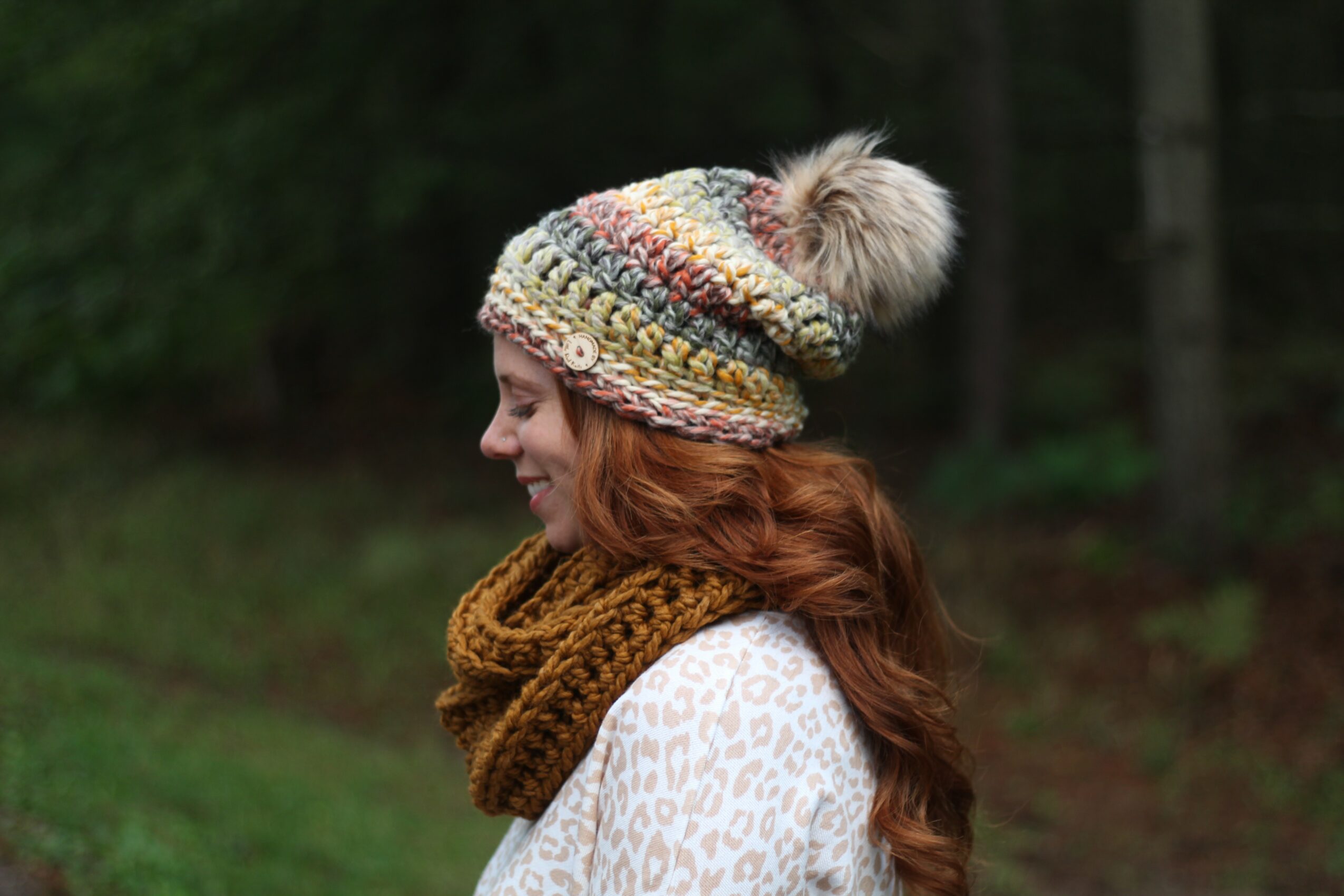 Woman with red hair wearing a colorful knit hat with pom-pom, a brown scarf, and a white patterned coat outdoors.