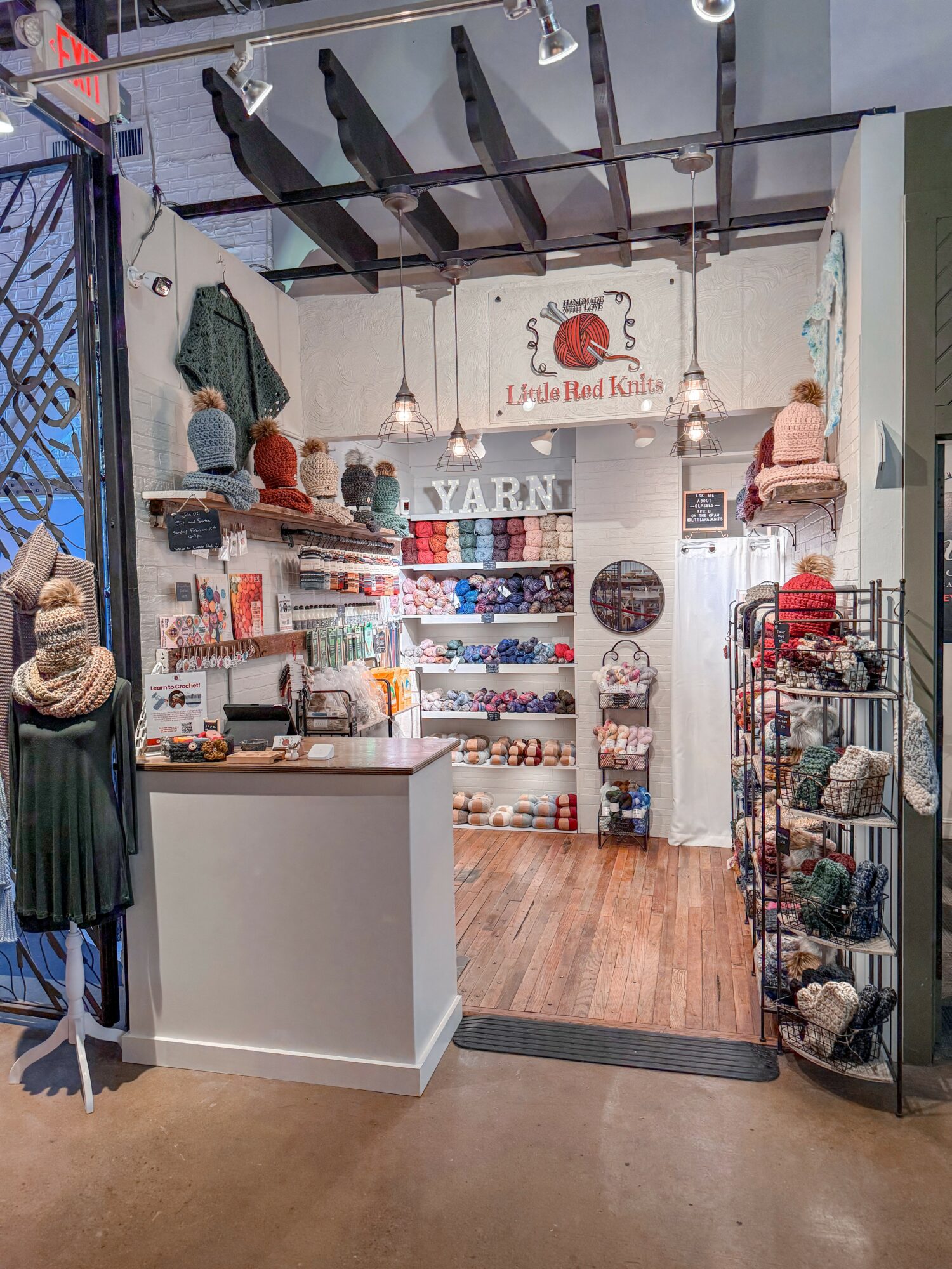 Store interior with shelves of yarn, hats, and accessories, and a counter at the front. Wooden floor and hanging lights.