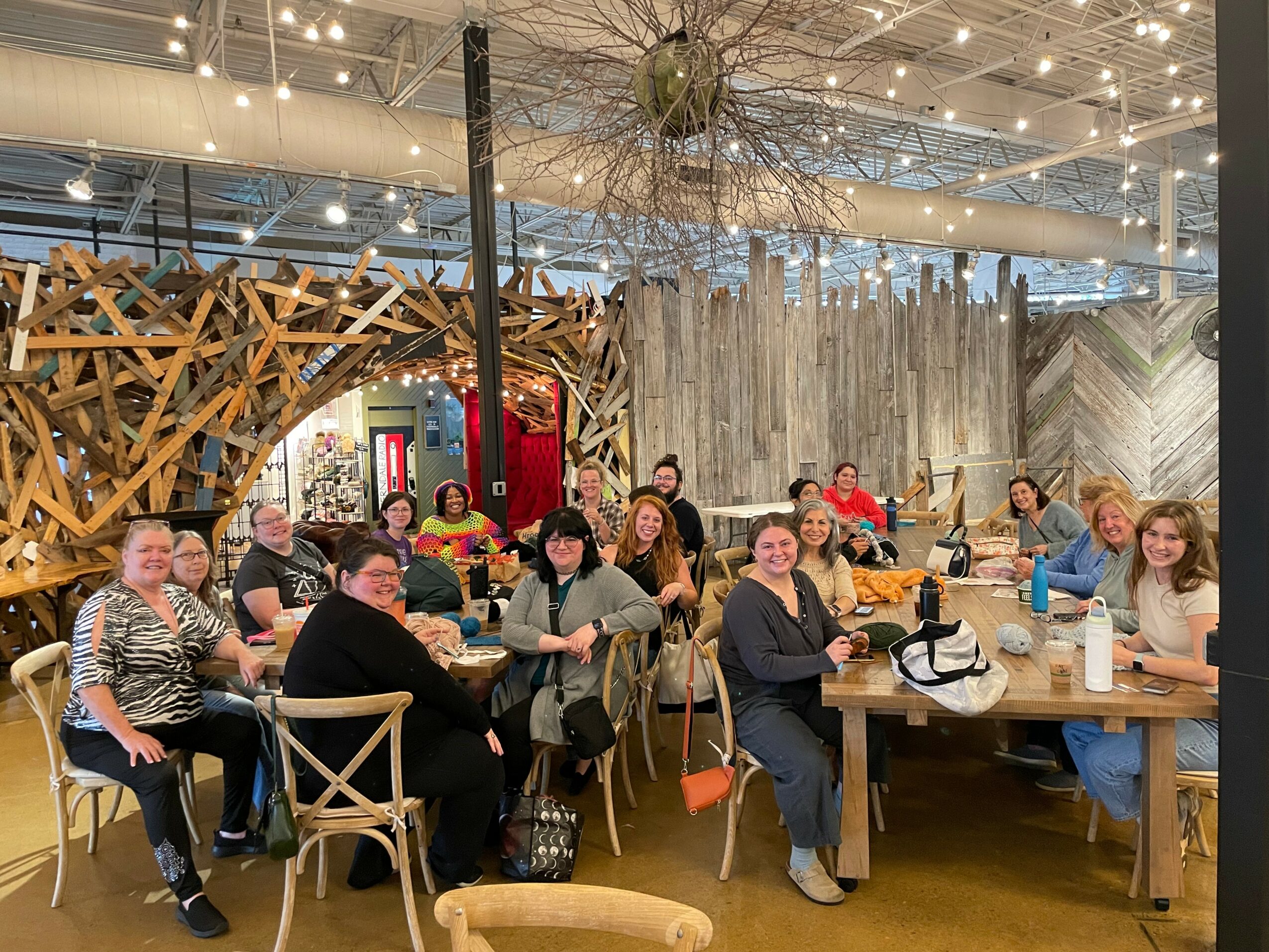 Group of people sitting at tables in a decorated indoor space with string lights and wooden structures.