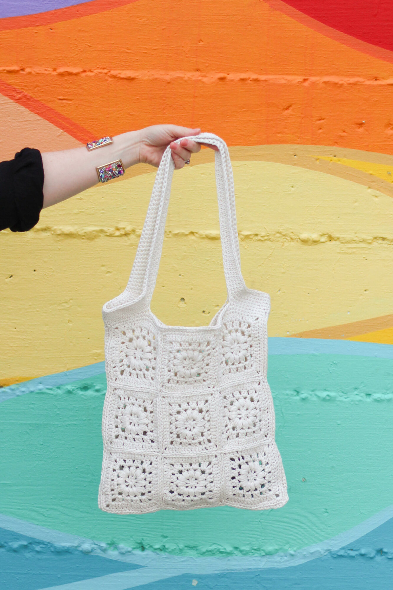 Person holding a white crocheted bag with a colorful painted background, arm extended to display the bag.