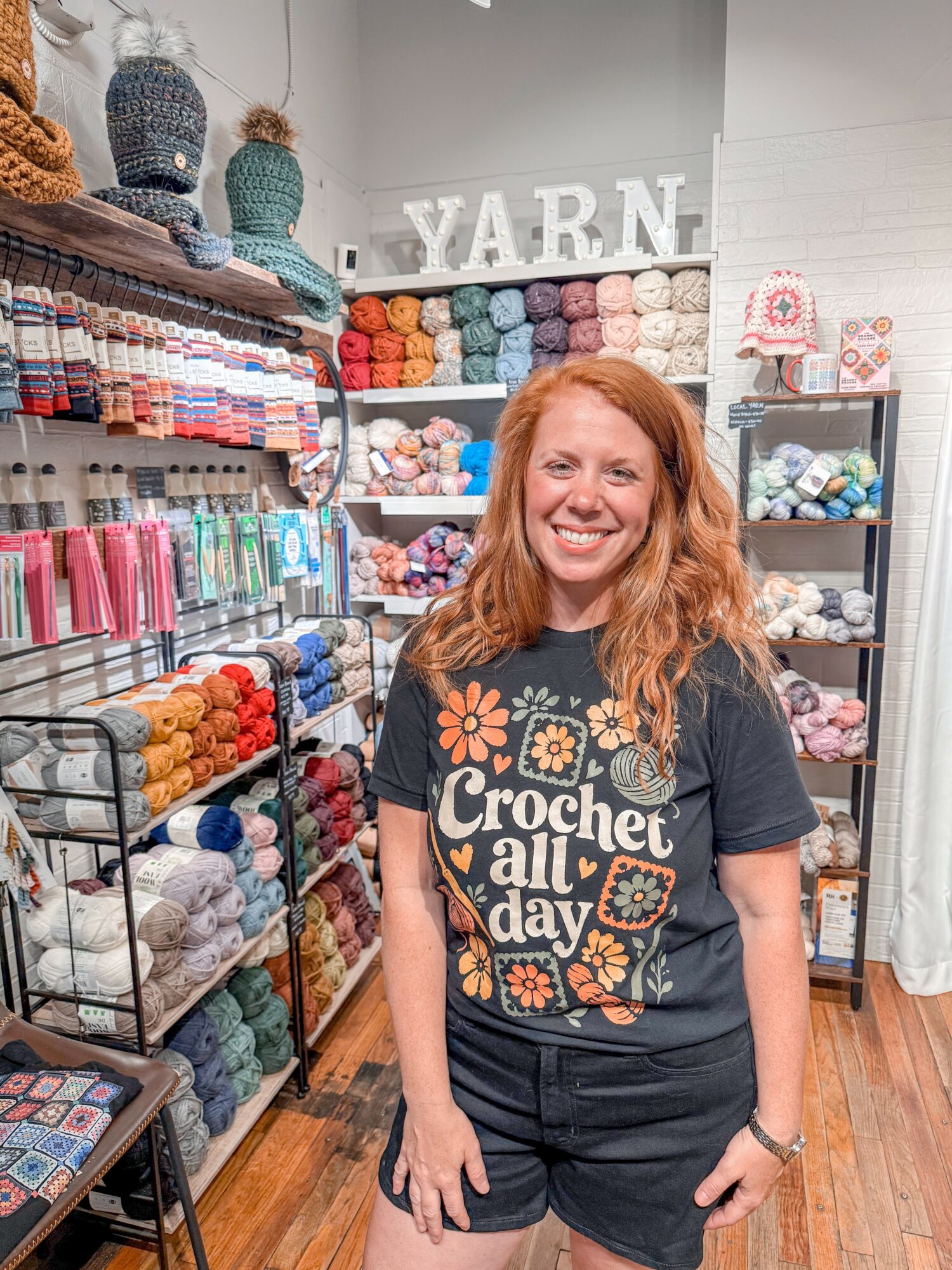 Smiling woman with red hair in a yarn store, surrounded by colorful yarn shelves and knitting supplies.
