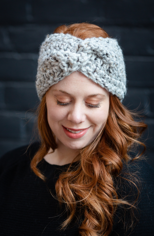 Woman with long wavy red hair wearing a chunky knit headband, smiling with eyes closed, dark background