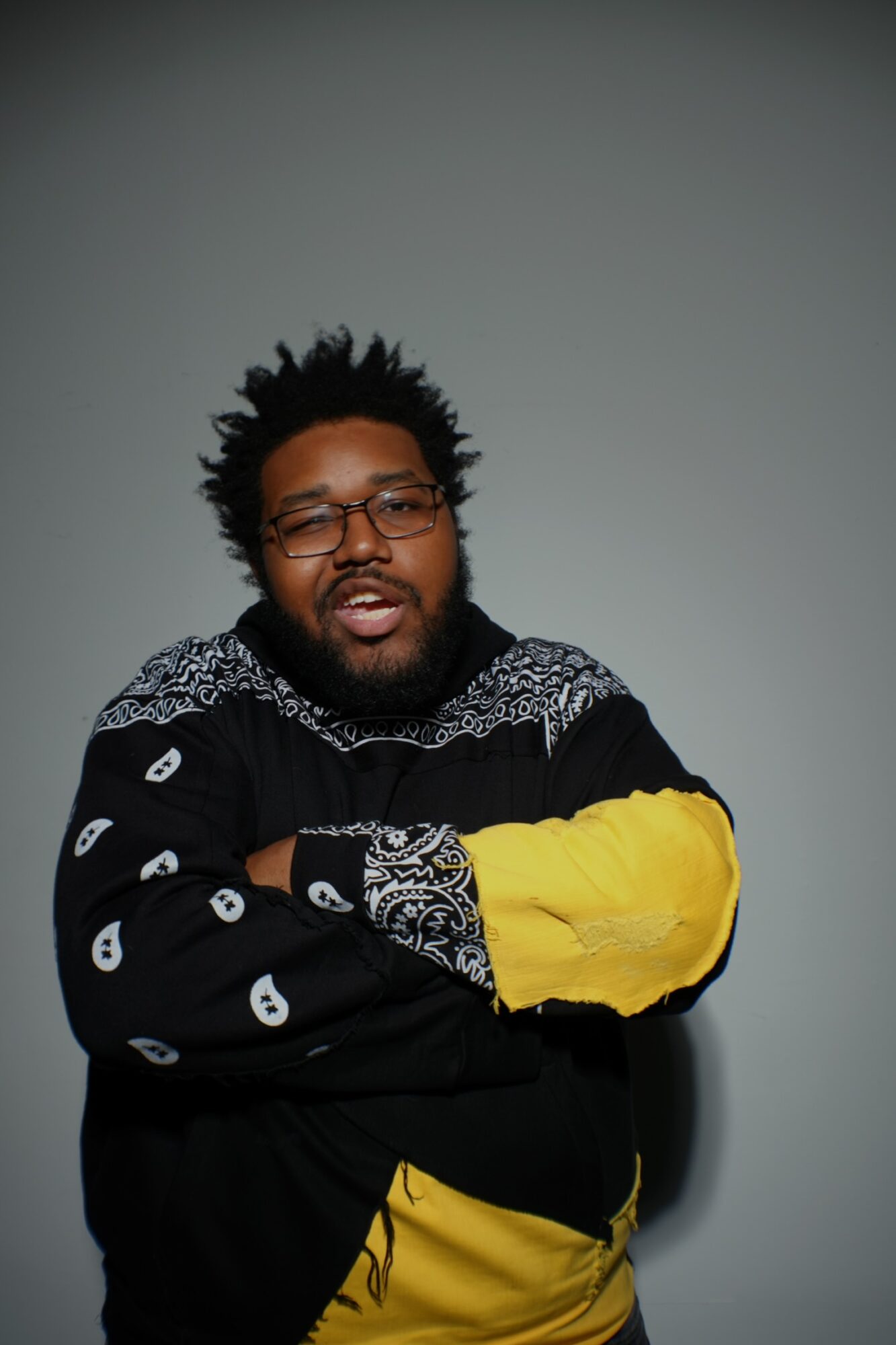 Man with glasses and a beard, wearing a black and yellow sweater, standing with arms crossed against a plain background.