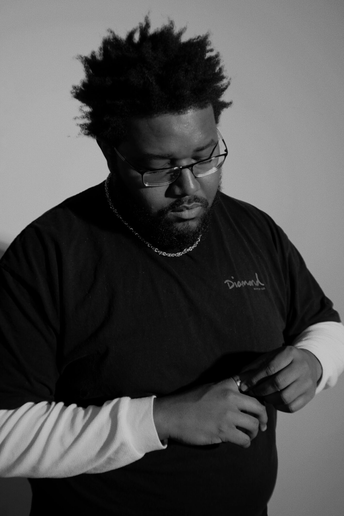 Person with glasses and textured hair looking down, wearing a black shirt and a chain necklace.