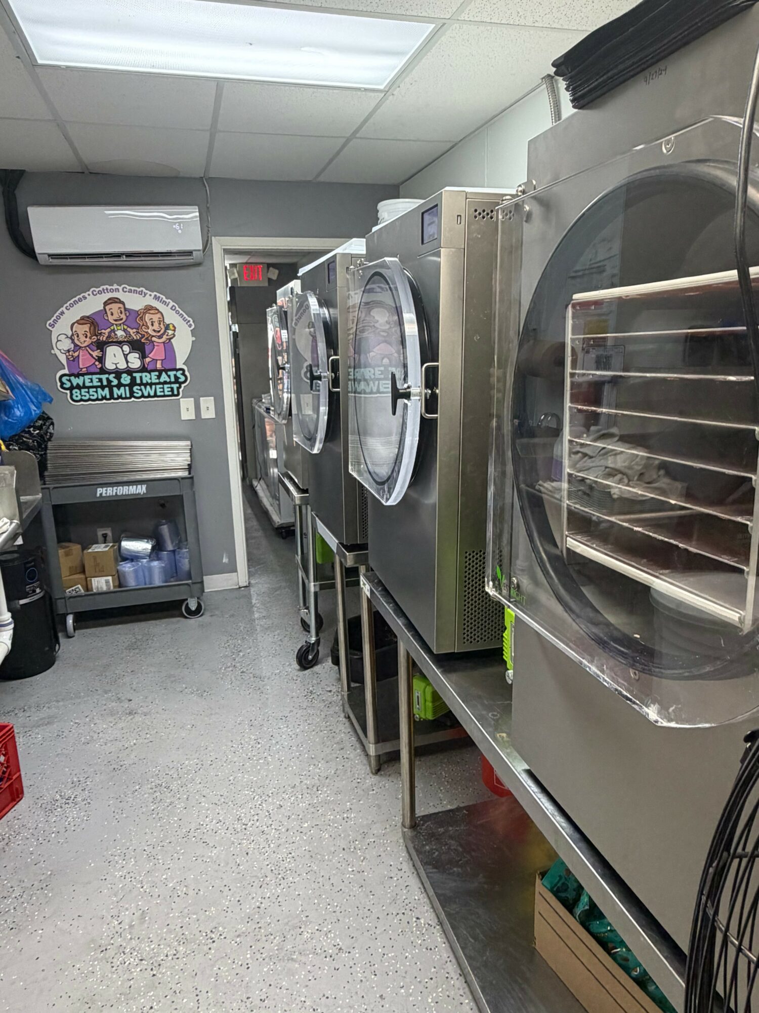Commercial laundry room with industrial washing machines and a cart, ceiling light, and a colorful sign on the wall.