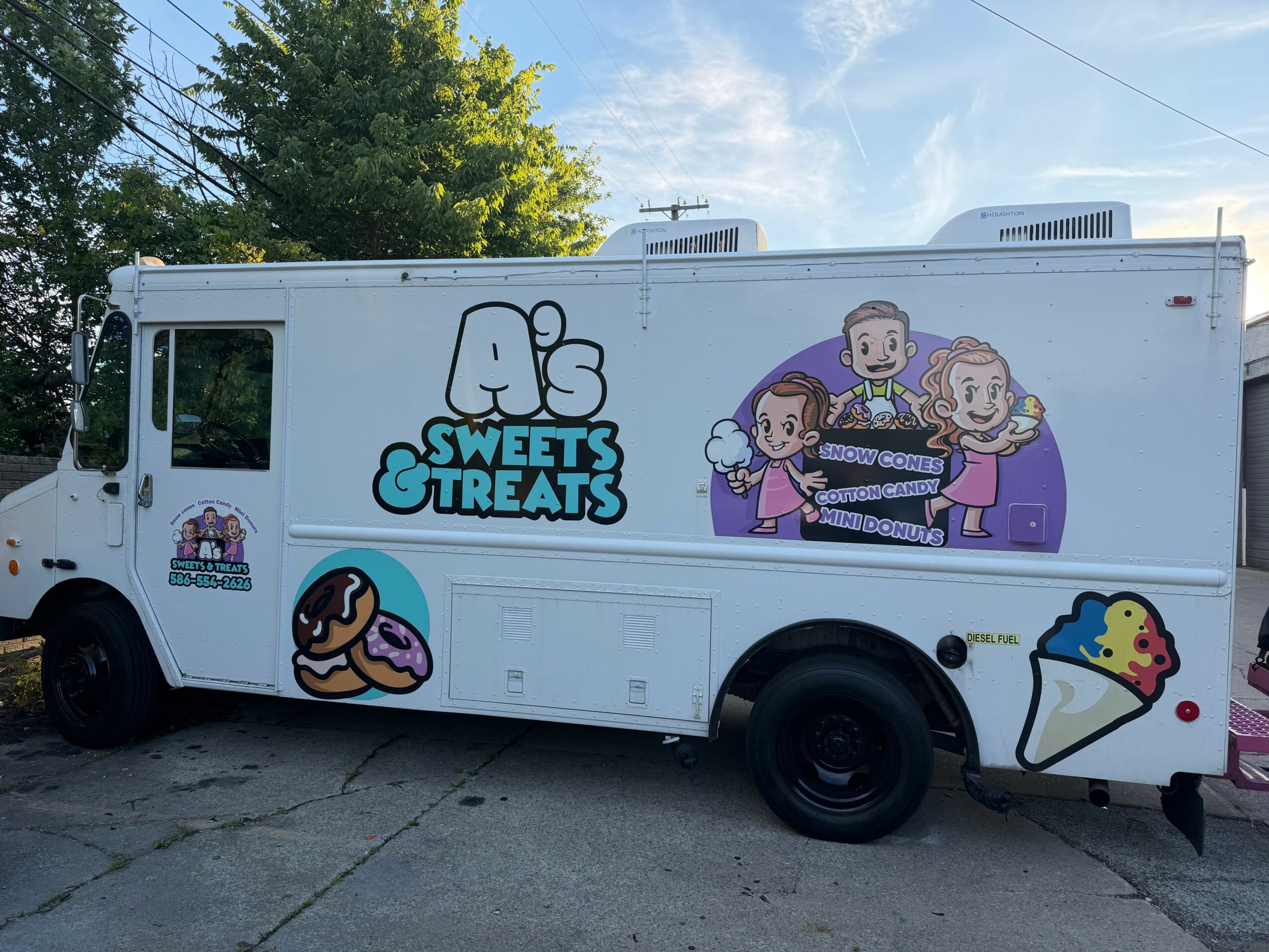 Ice cream truck with cartoon characters and text, parked outdoors with trees and sky in background.