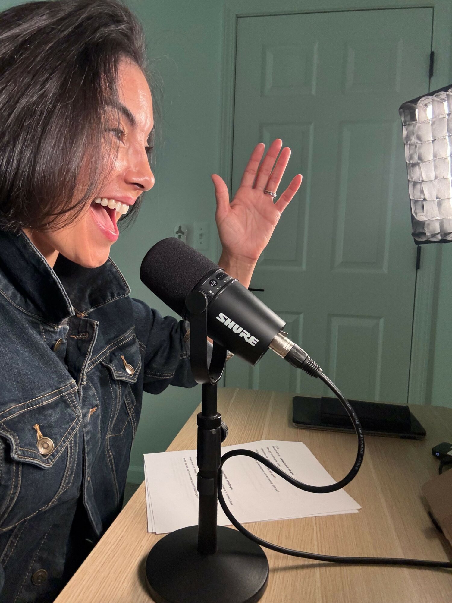 Woman speaking into a microphone, gesturing with her hand, sitting at a desk with papers and a computer mouse.