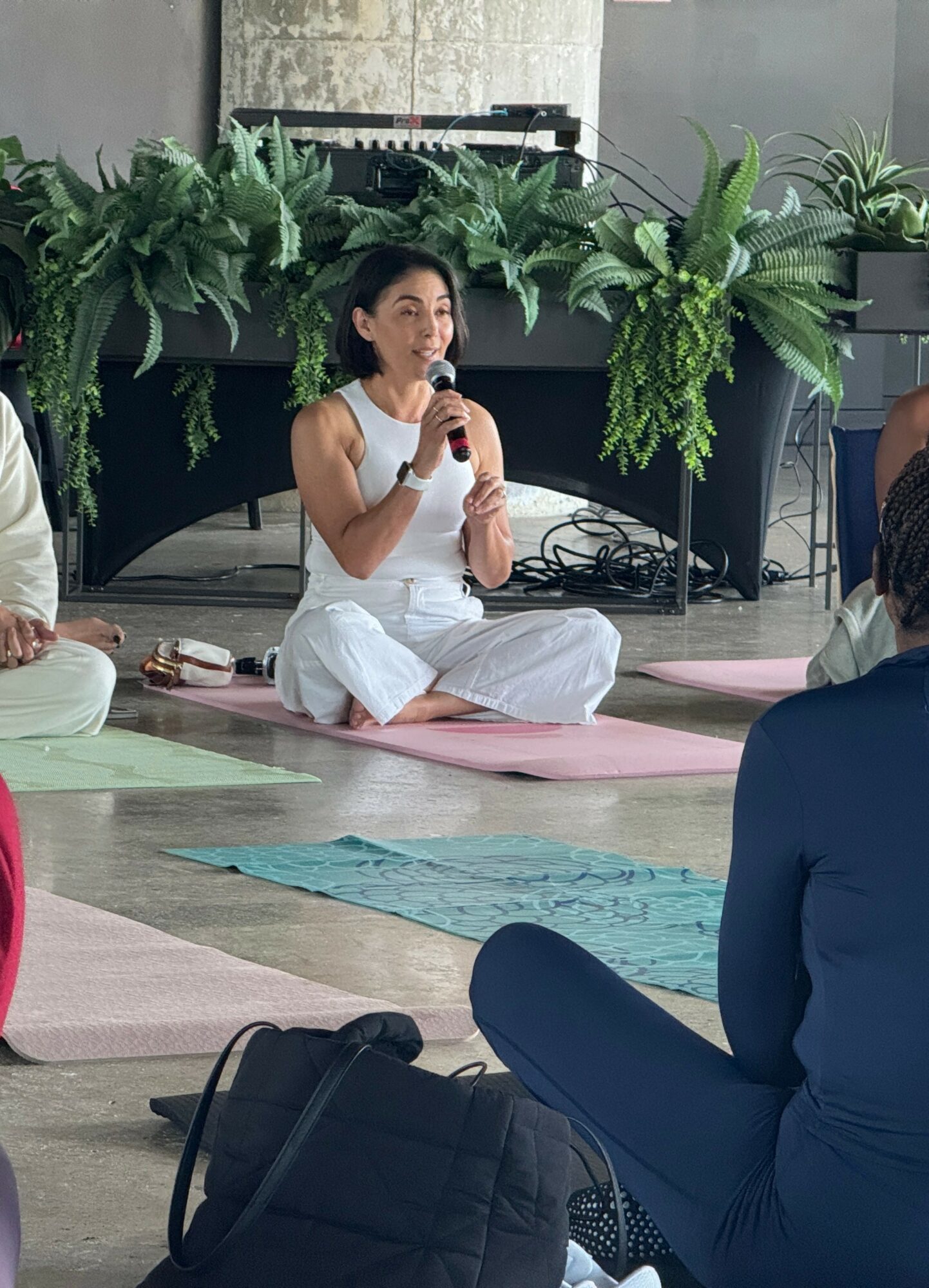 Woman sitting cross-legged on a yoga mat, speaking into a microphone, surrounded by plants and people, indoors.