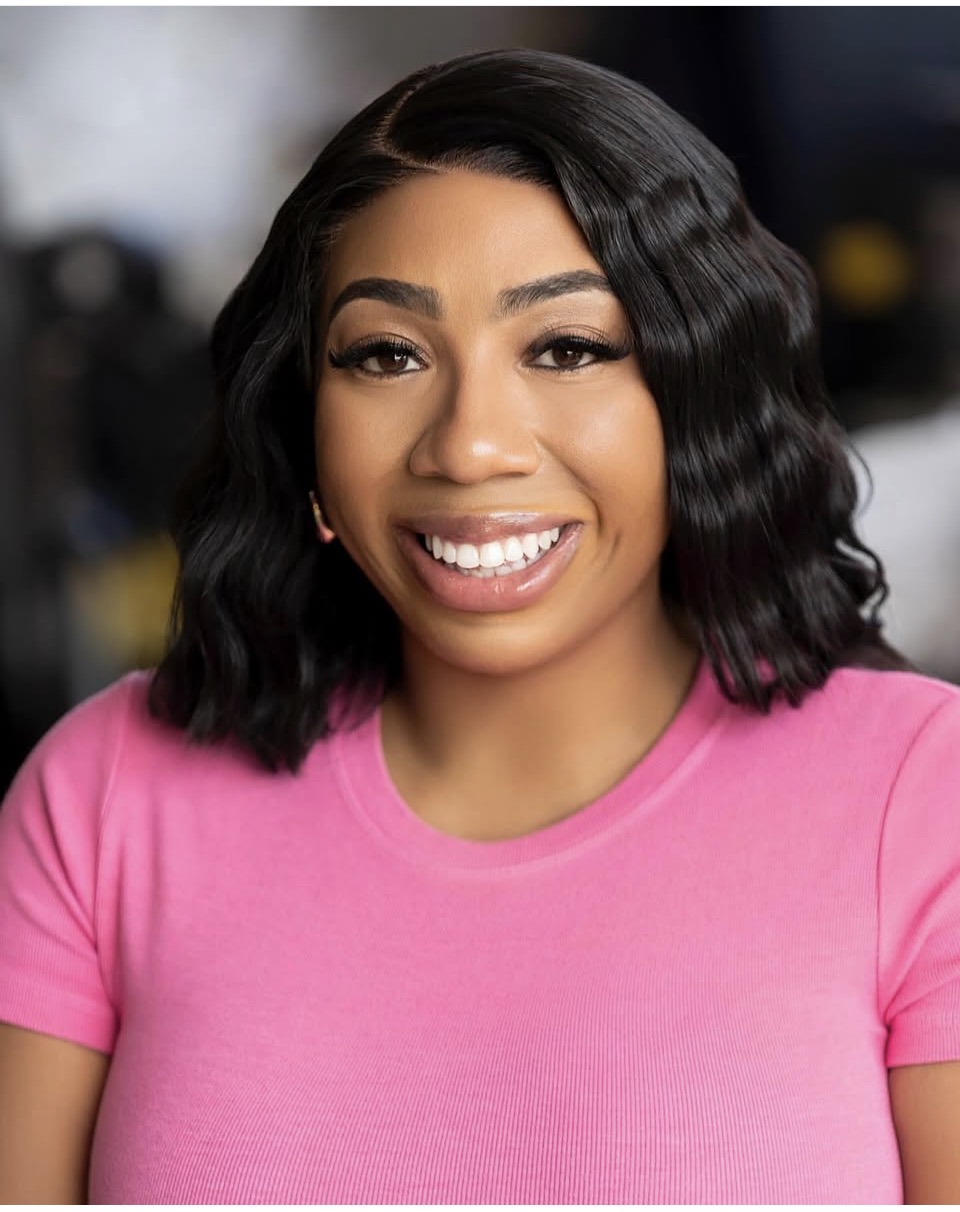 Young woman with wavy black hair smiling, wearing a pink shirt, in an indoor setting.