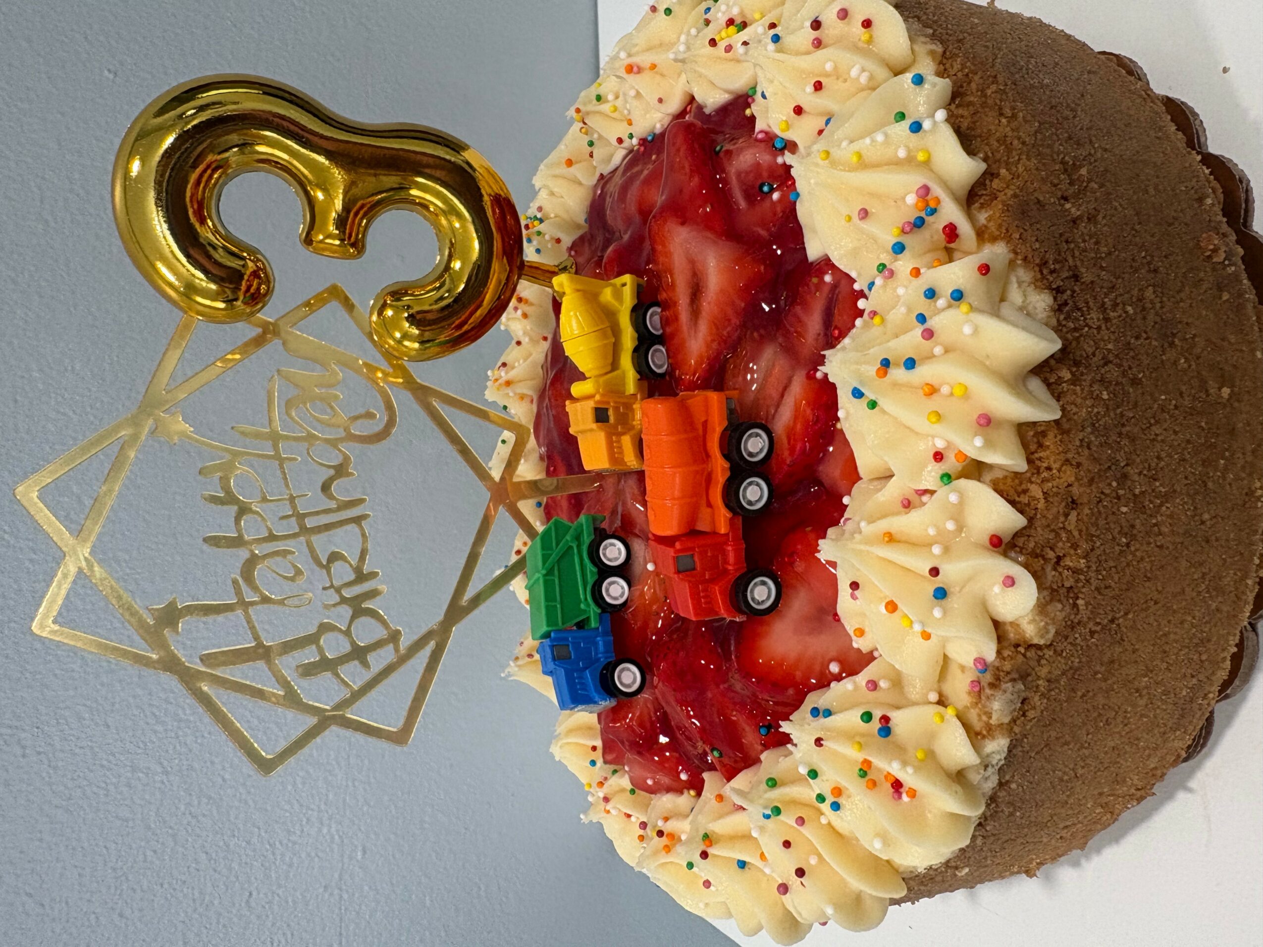 Chocolate birthday cake with whipped cream, strawberries, and colorful sprinkles, decorated with a gold number 3 candle and a gold 'Happy Birthday' topper.