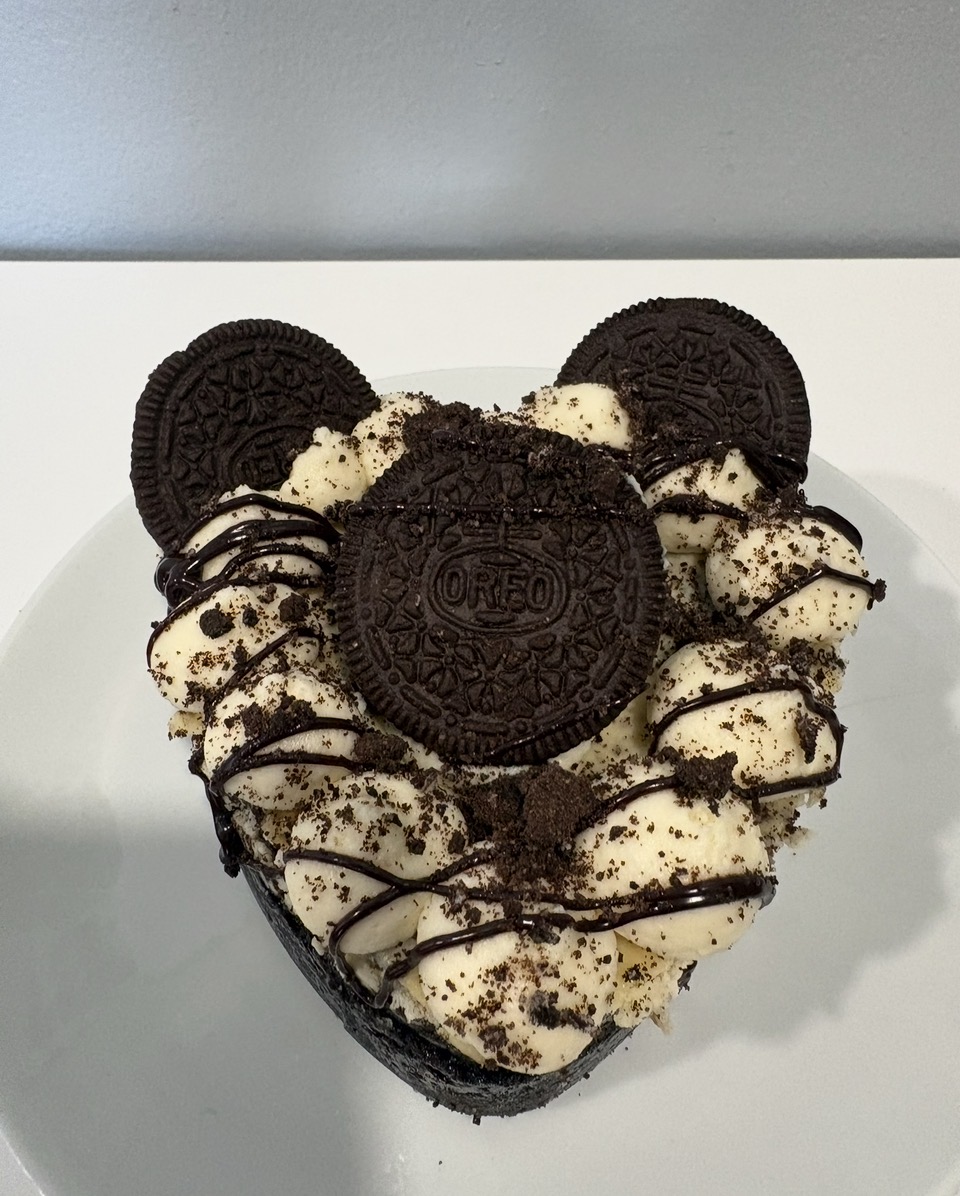 Heart-shaped cake decorated with Oreo cookies, cream, and chocolate drizzle, on a white surface.