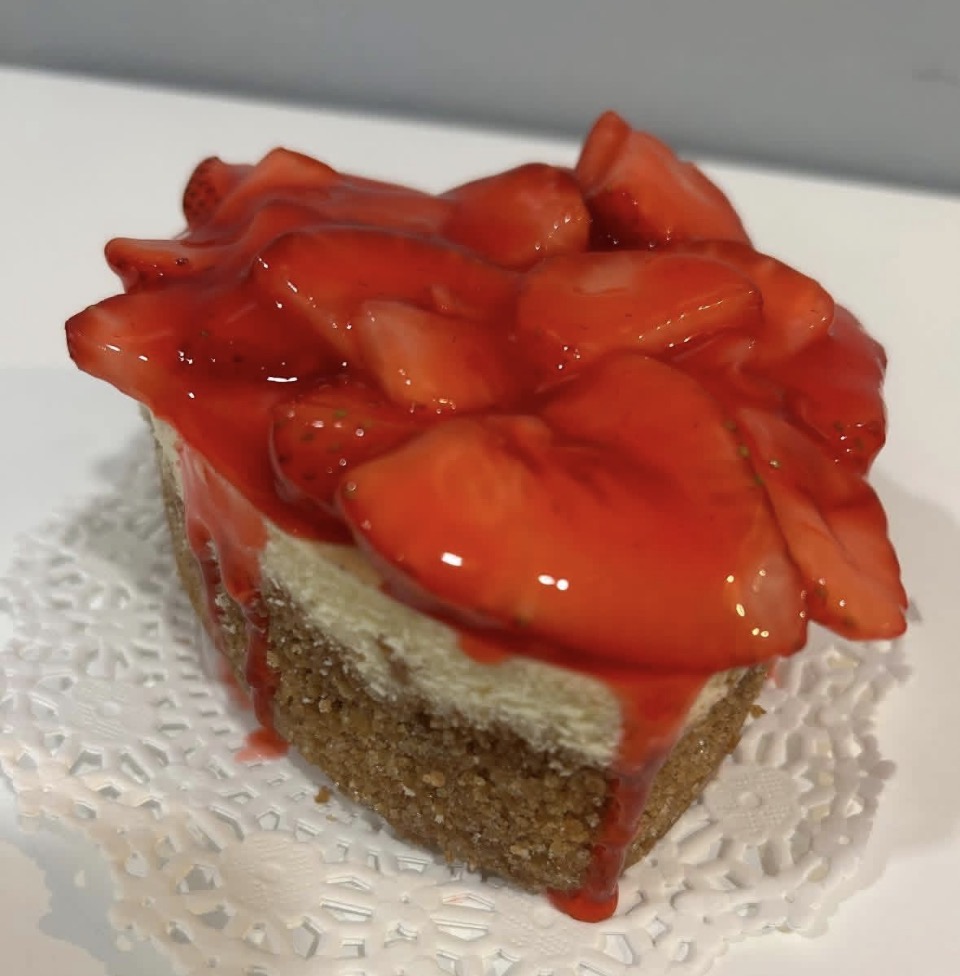 Slice of strawberry cake with strawberries and red glaze on top, on a doily, against a plain background.