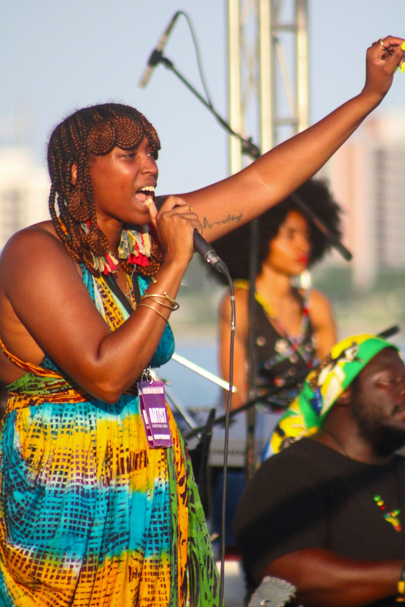 Woman singing into microphone with arm raised, wearing colorful dress, outdoors during daytime, with other performers in background.