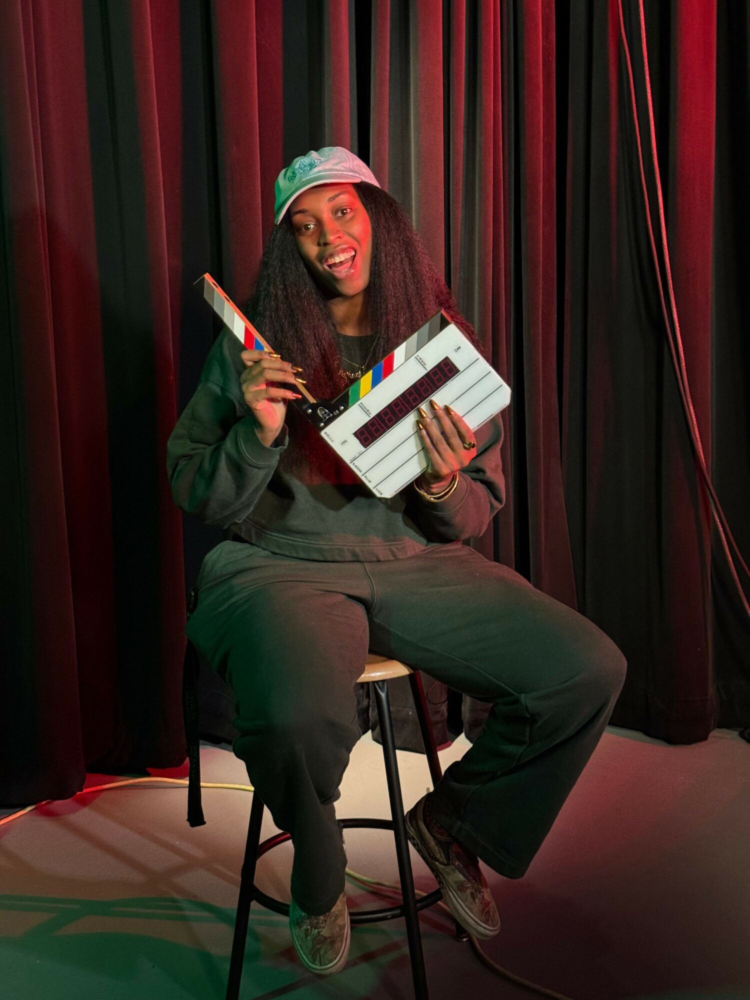Person sitting on a chair holding a clapperboard in front of a dark curtain, wearing a green headband and dark clothing.