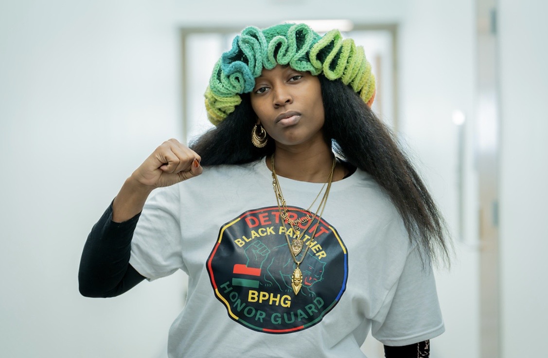Young woman with colorful hair styled in large curls, wearing a gray T-shirt with a graphic design, standing indoors.