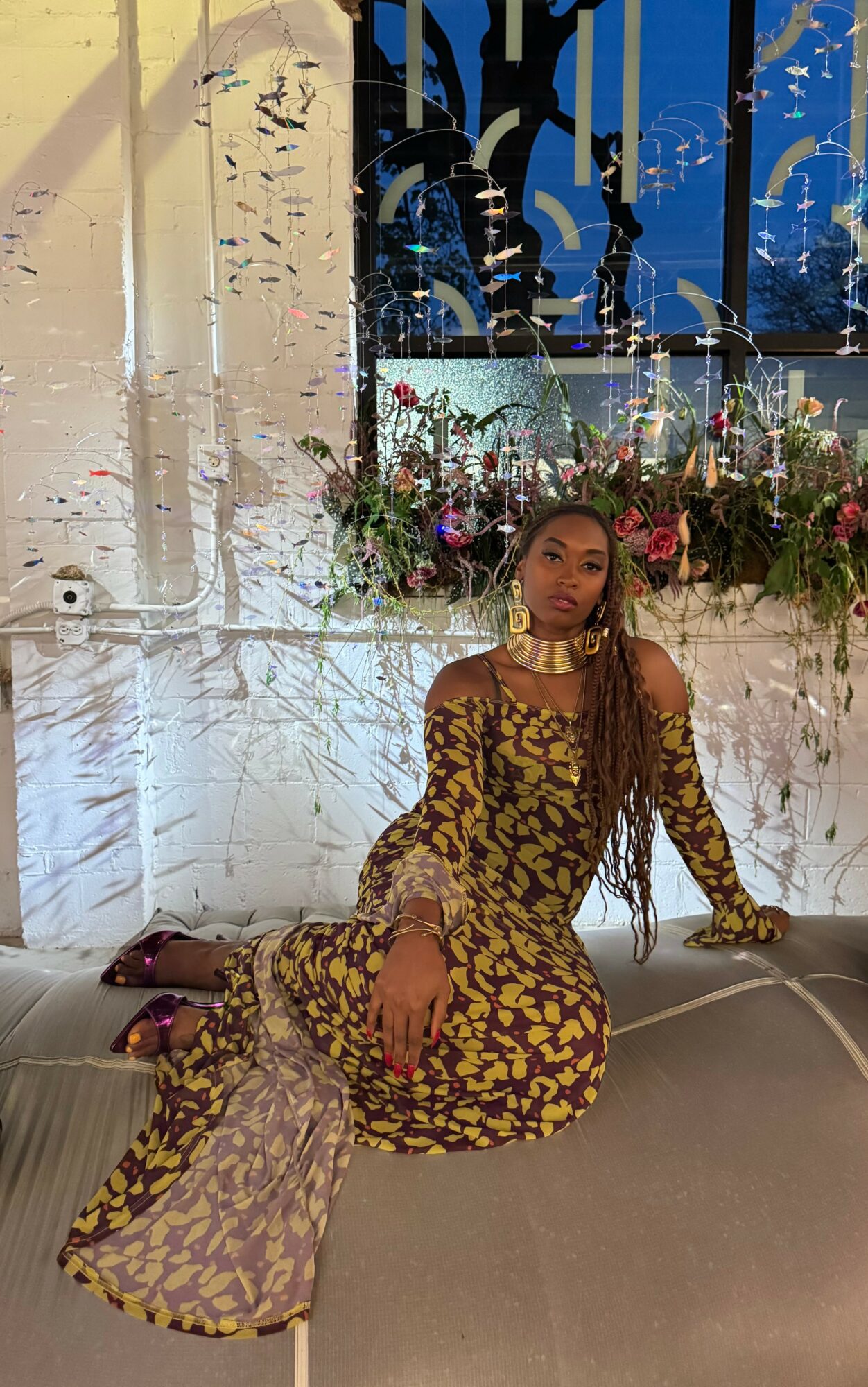 Woman in yellow and black patterned dress sitting on the floor with floral backdrop behind her.