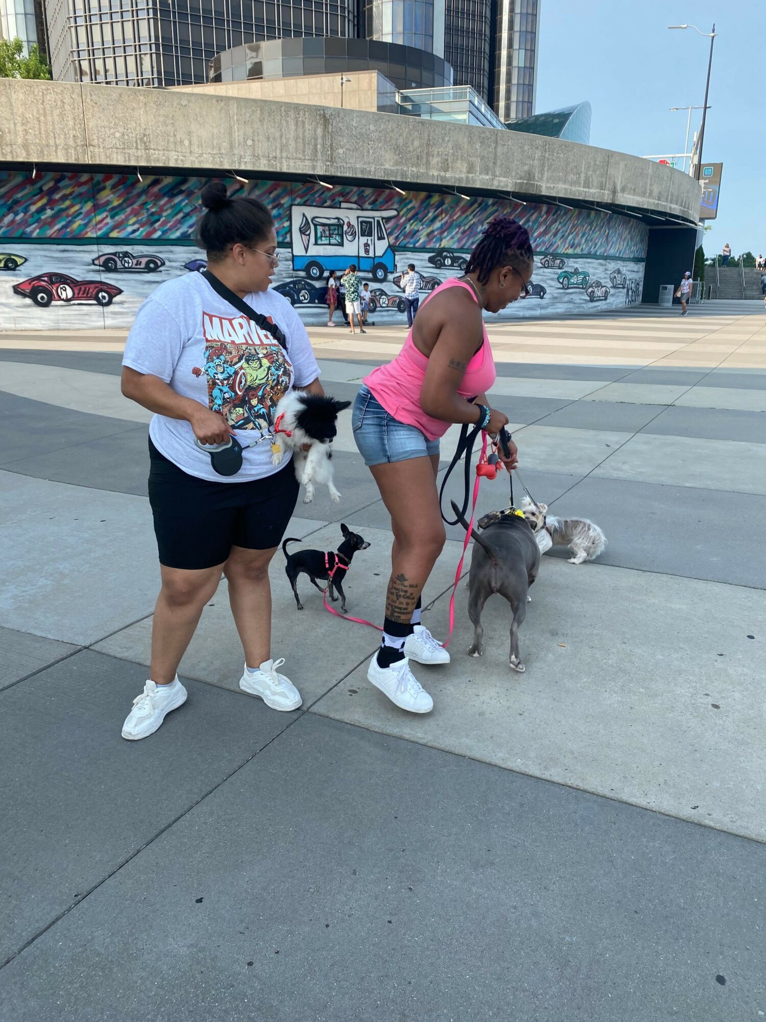 Two women walking dogs on a city sidewalk, with a mural and parked cars in the background.