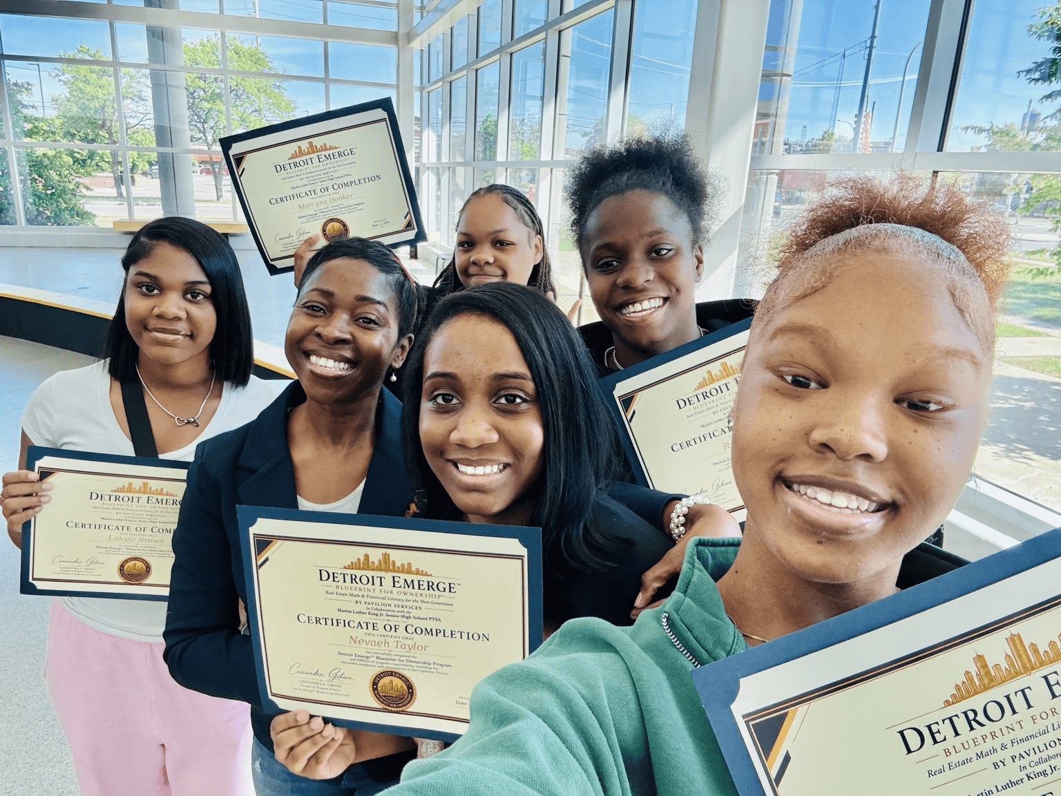 Six young women smiling and holding certificates in a bright room with large windows.
