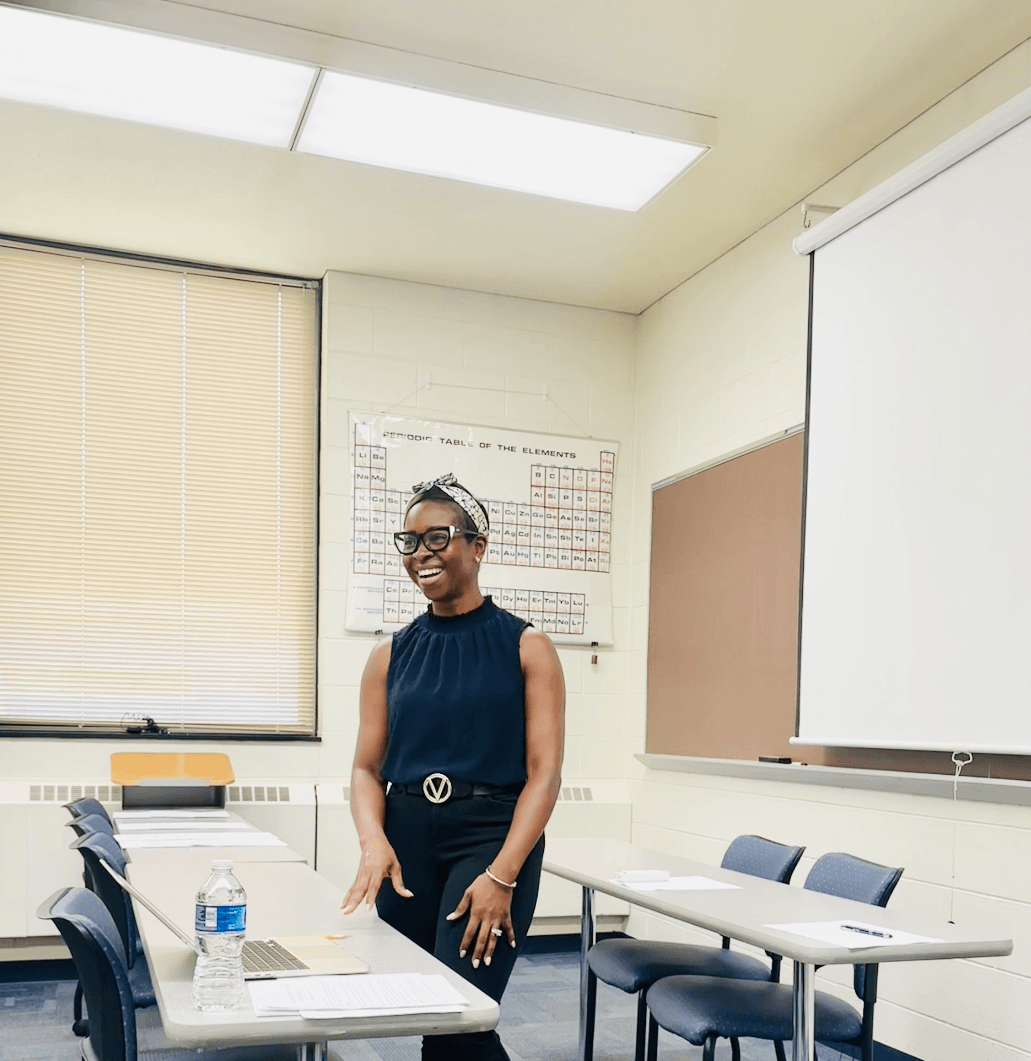 Woman standing in a classroom, smiling, wearing glasses and a dark sleeveless top, with desks and a whiteboard behind her.