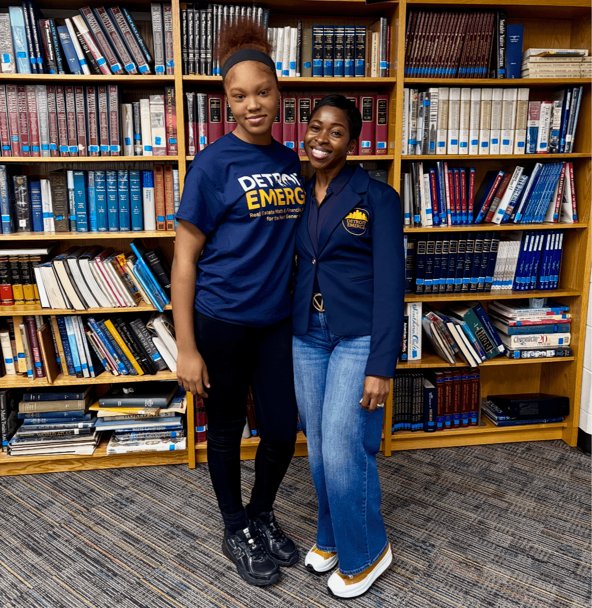 Two women stand in front of a bookshelf filled with books, smiling and posing together.
