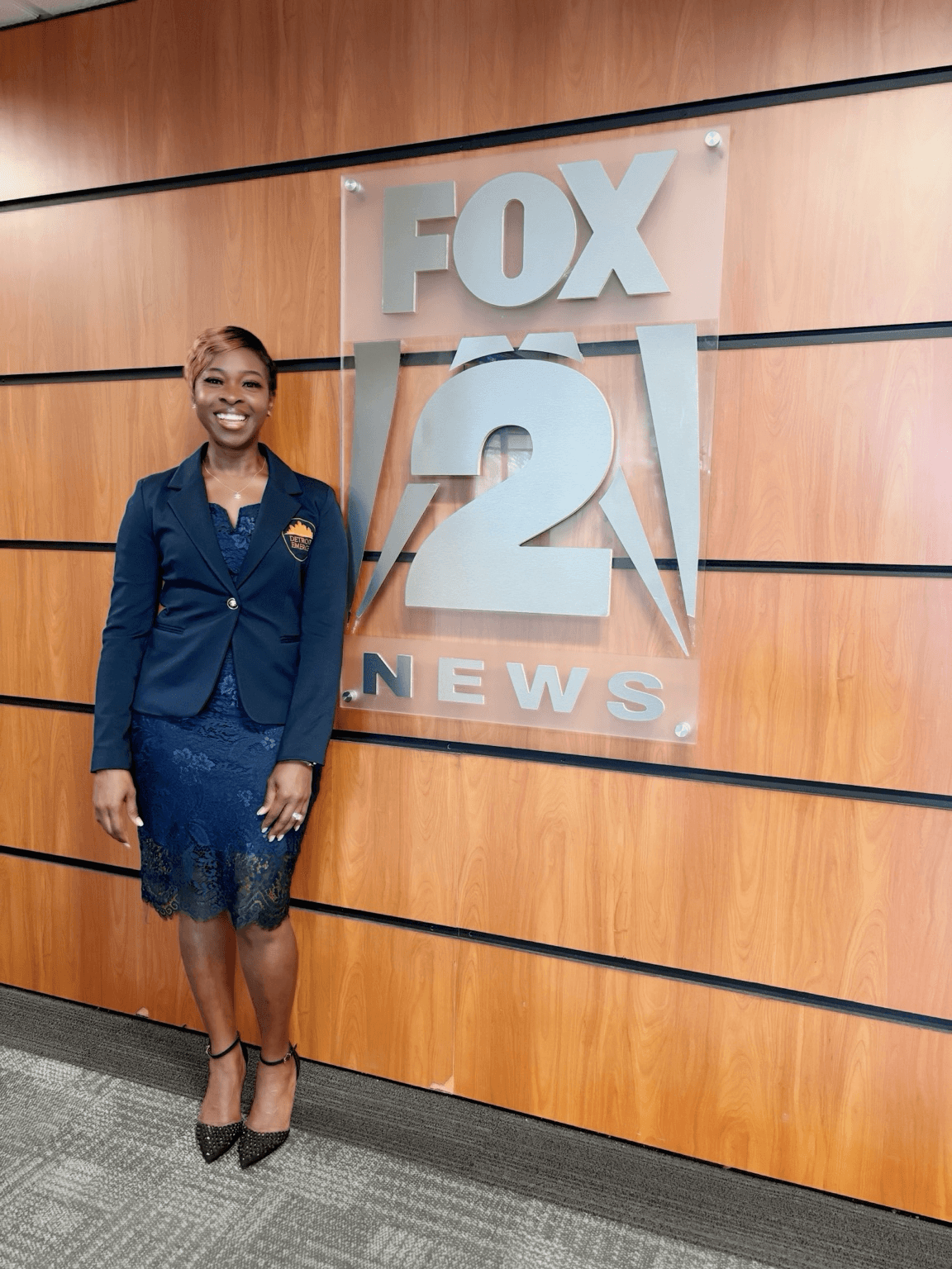 Woman in professional attire standing in front of FOX 2 News logo on wooden wall.