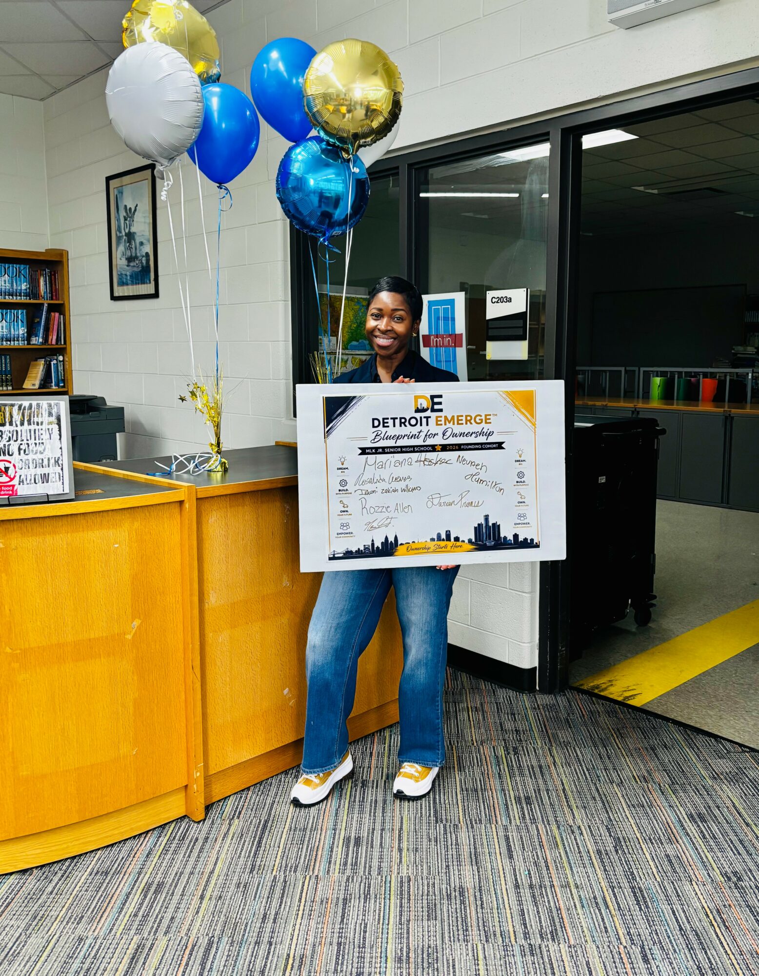 Person holding a large check with balloons behind them, standing in an indoor space with a yellow counter and bookshelves.