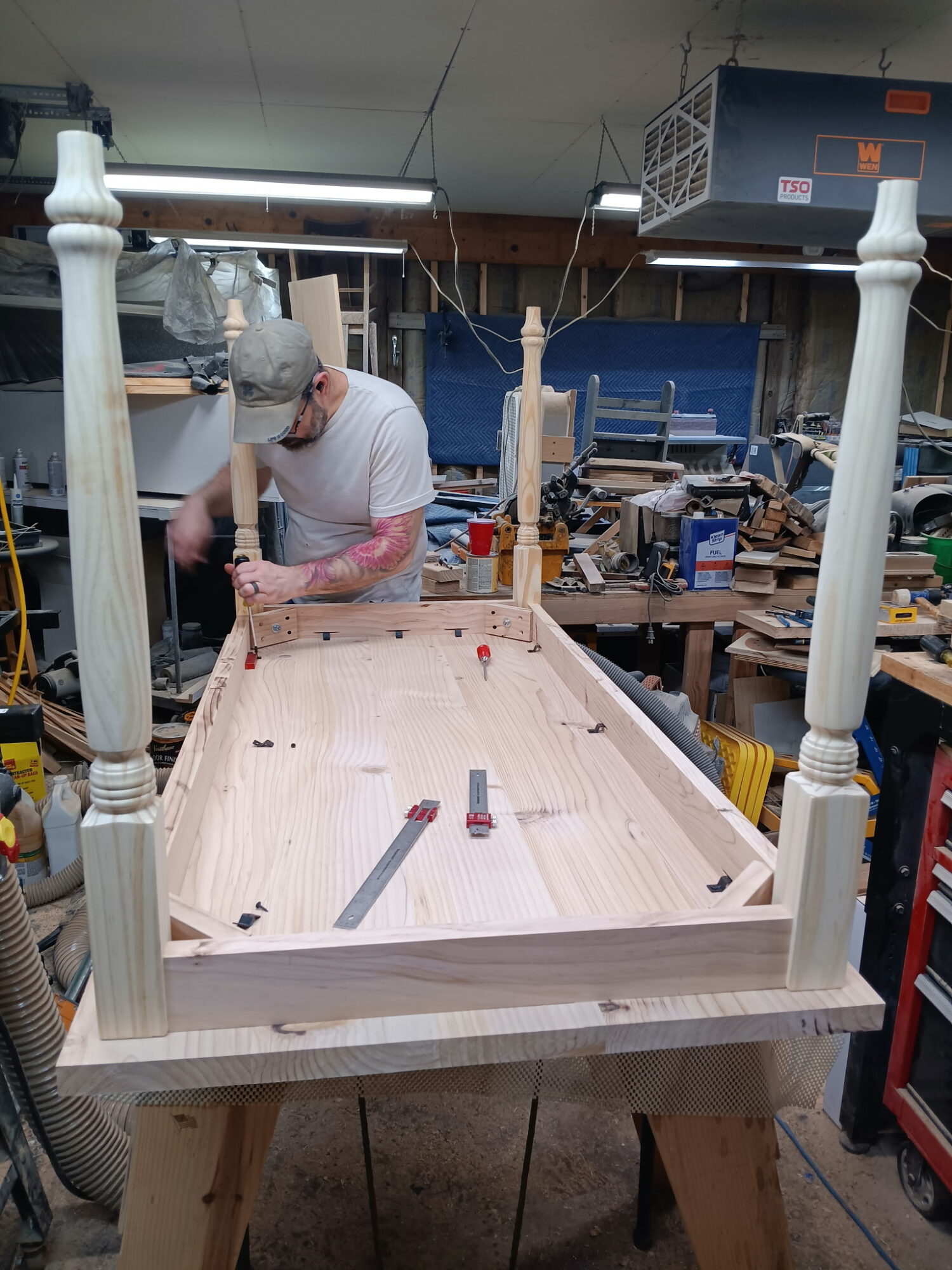 Person working on a wooden bed frame in a workshop, surrounded by tools and materials.