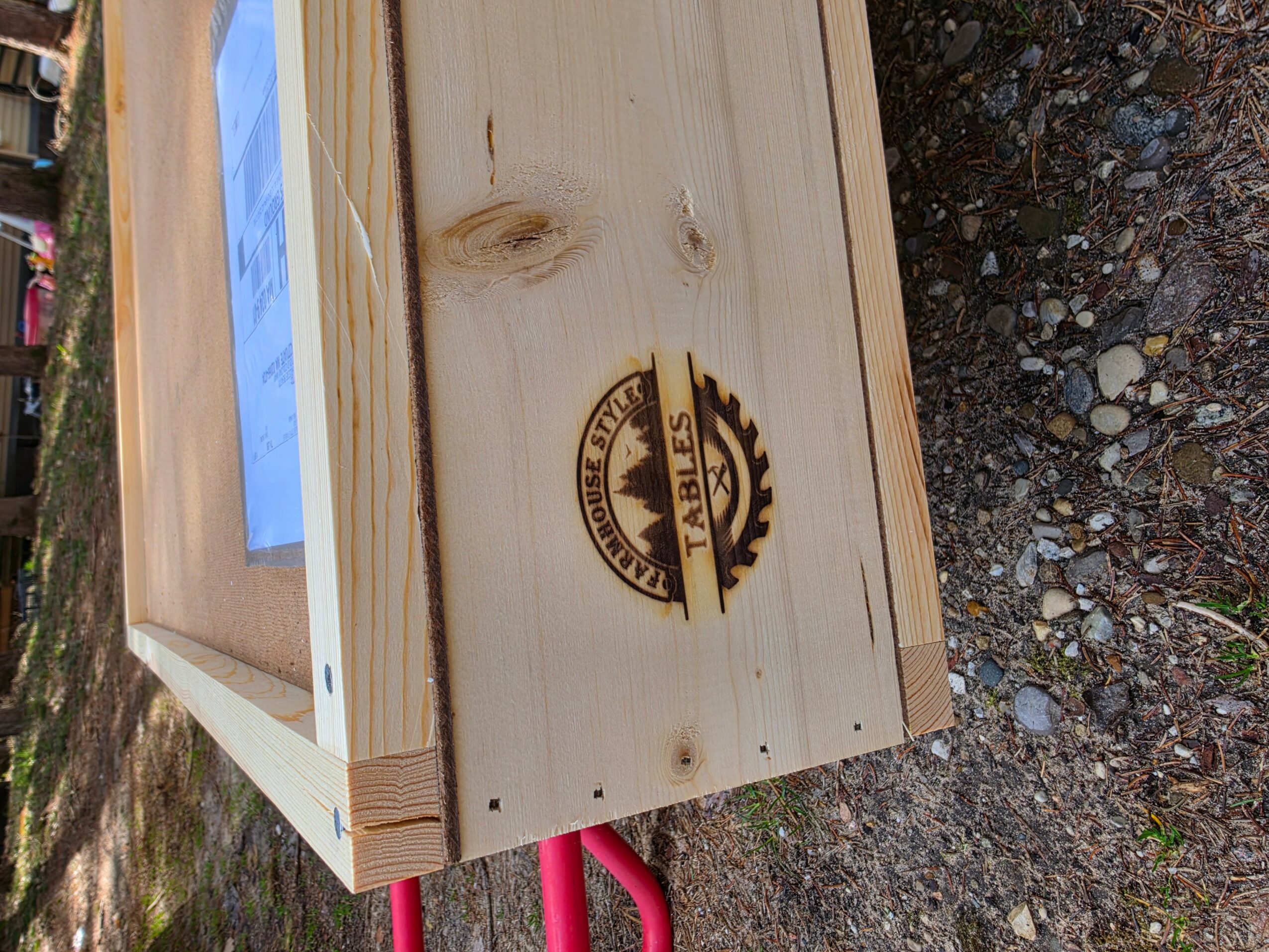 Wooden box with a logo on the side, outdoors on gravel, with a blue paper or sign inside.