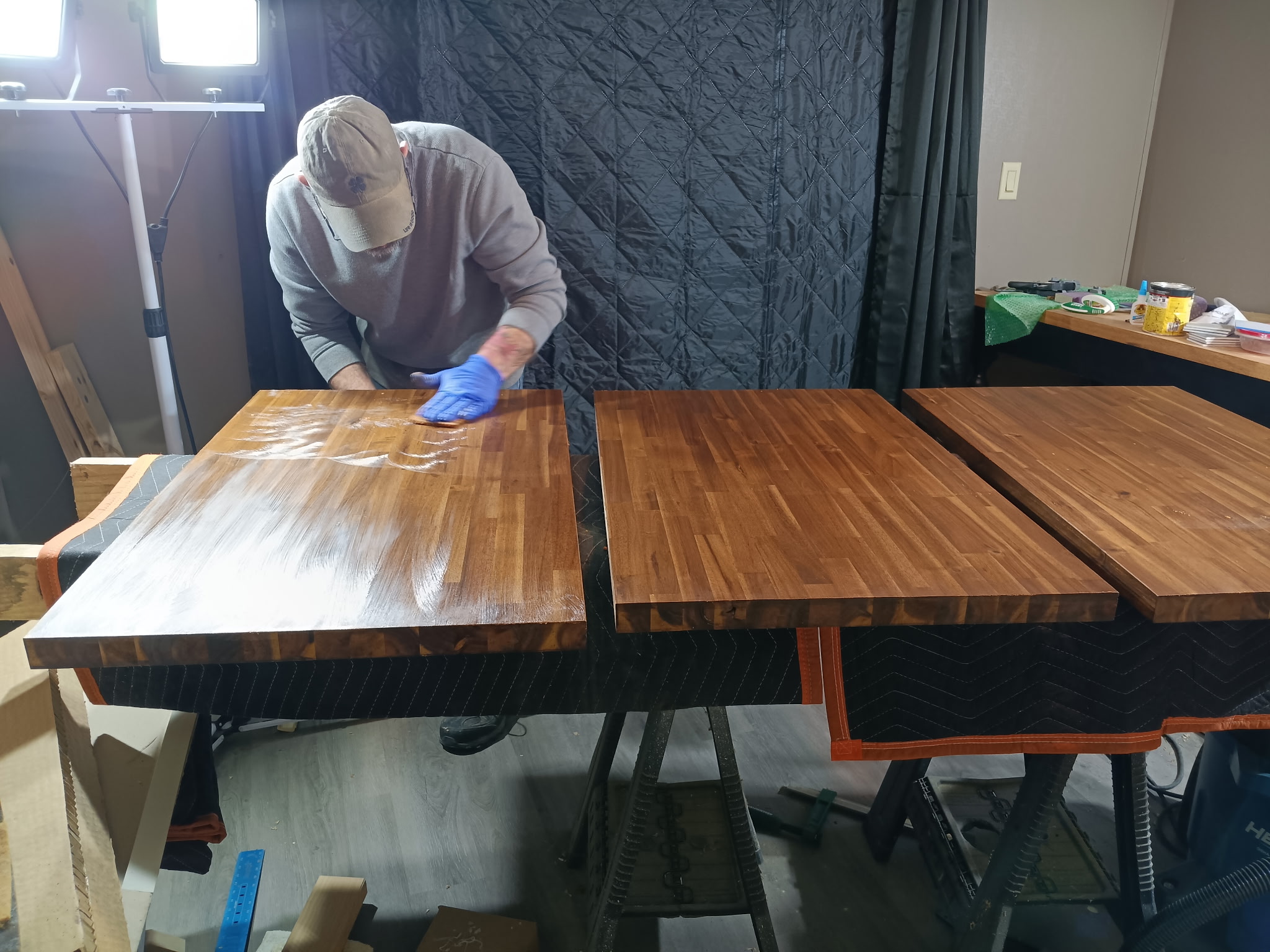 Person wearing a cap and gloves working on a wooden table, with other tables nearby in a room.