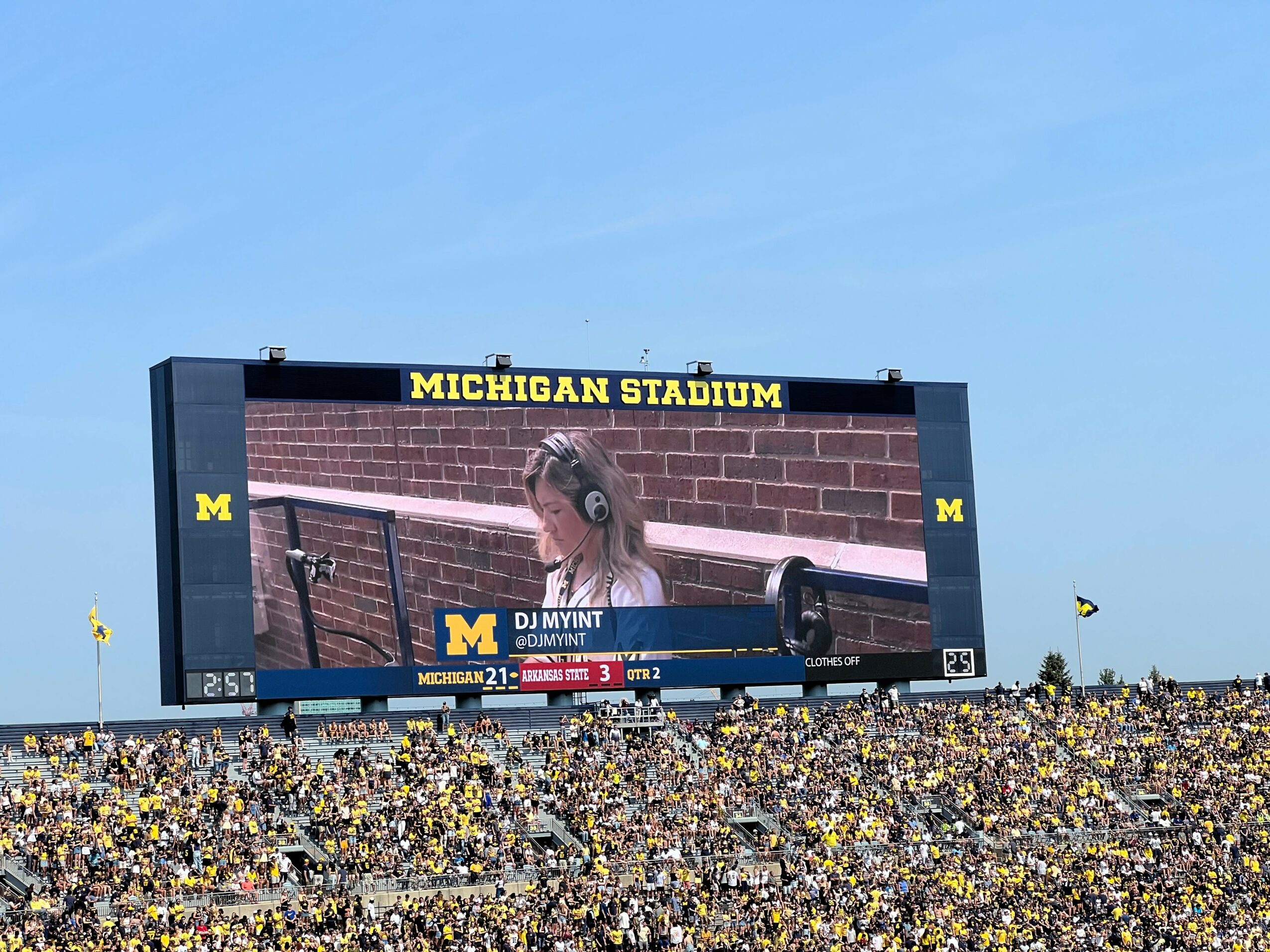 Large outdoor stadium scoreboard displaying a person with headphones, with a crowd below and a blue sky above.