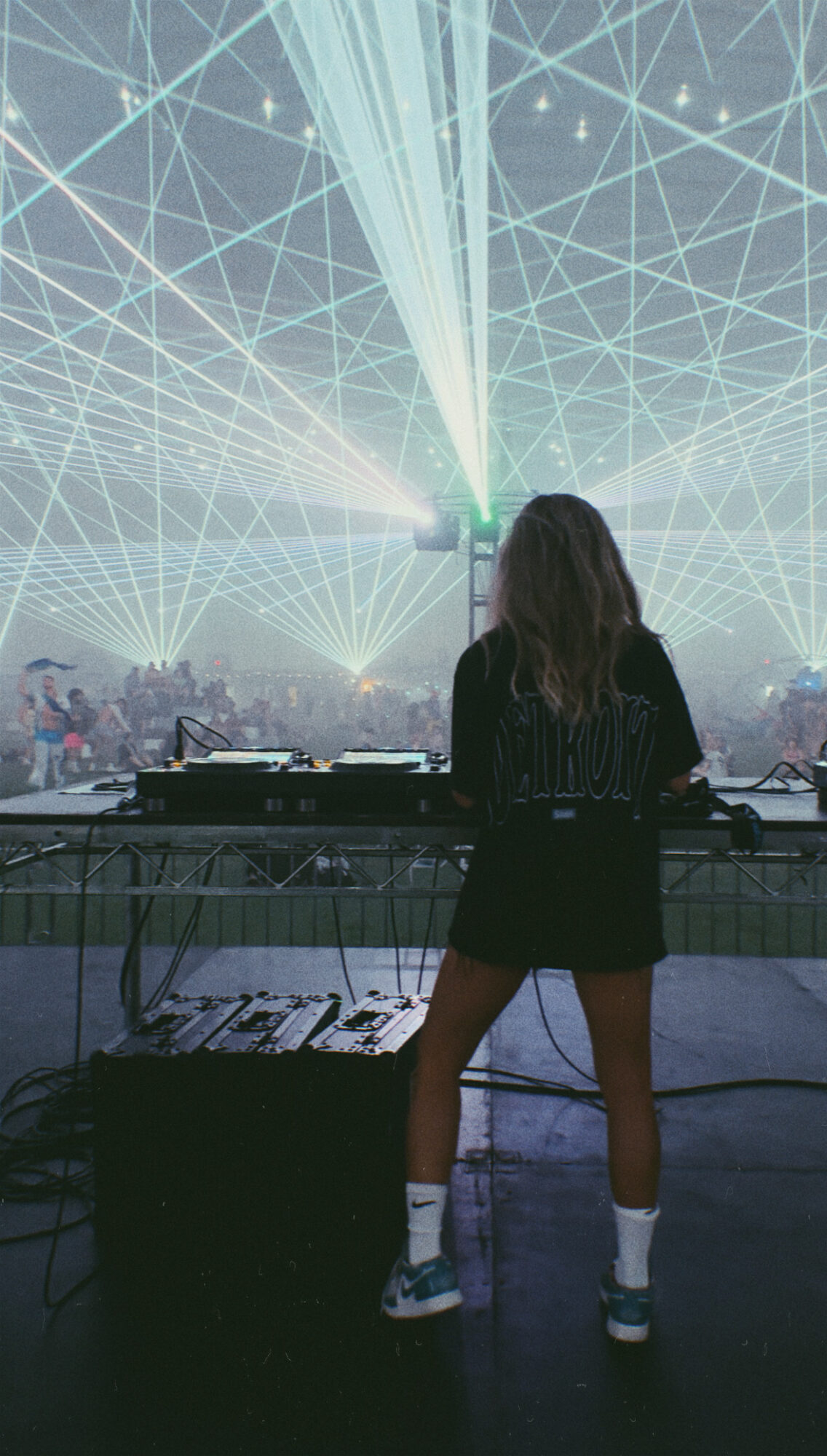 Person with long hair standing at DJ booth with laser lights and crowd in background.