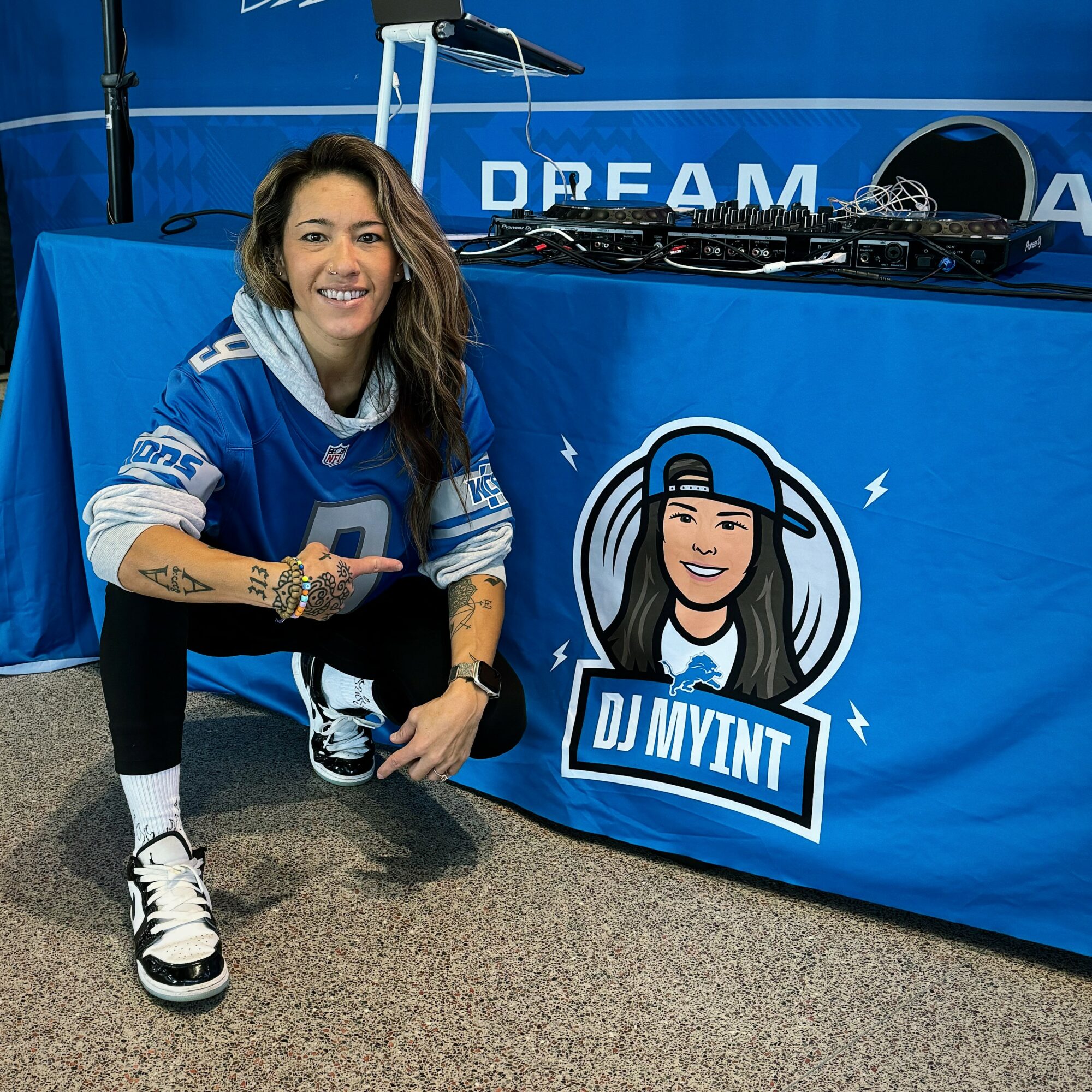 Woman crouching next to DJ booth with DJ MYINT logo, smiling, wearing a blue sports jersey, sneakers, and tattoos visible on arm.
