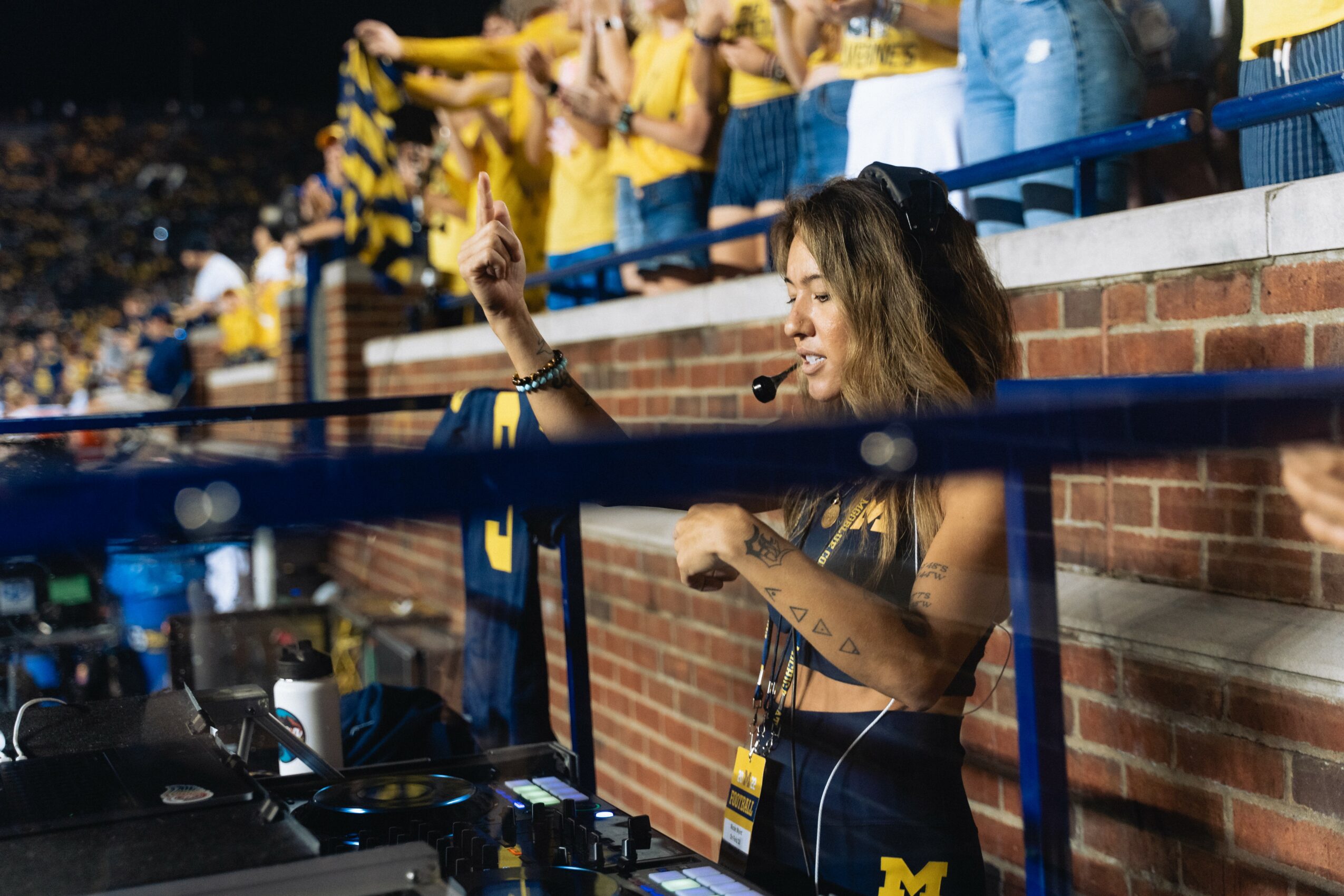 Woman with a headset and microphone, gesturing with her right hand, standing behind a barrier at a sports event.