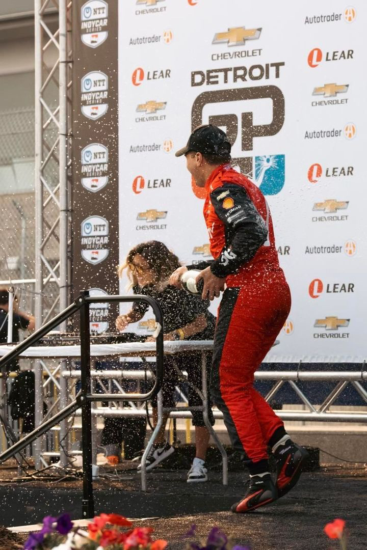 Two race car drivers celebrate on stage, one spraying champagne, with a backdrop of sponsor logos and a metal railing.